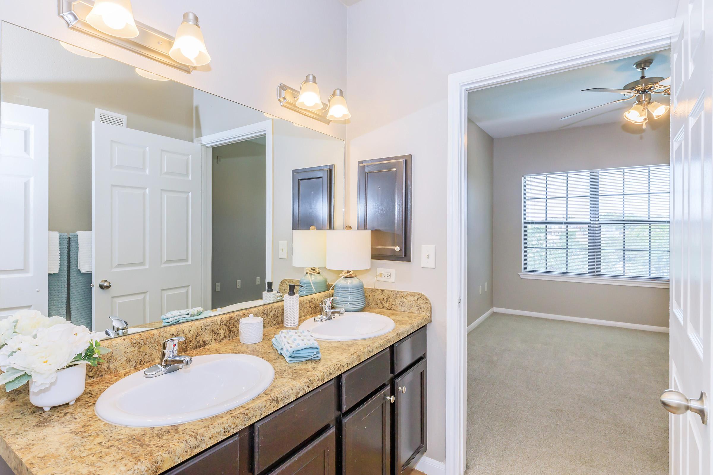 A well-lit bathroom featuring a double sink with granite countertop, polished fixtures, a stylish lamp, and decorative elements like flowers. A doorway leads to a cozy bedroom with light-colored walls and a ceiling fan, and a window providing natural light.