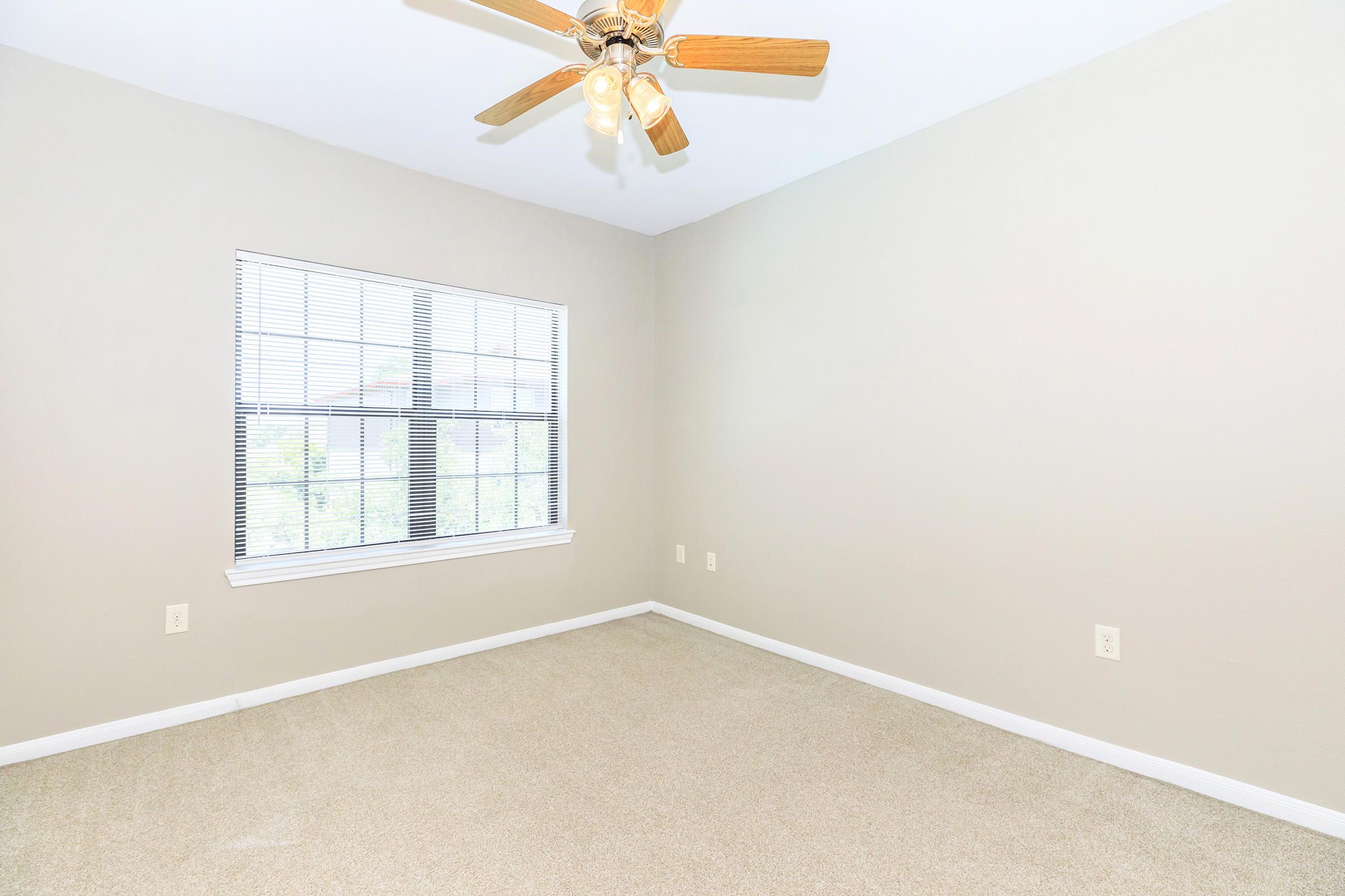 Empty room with light beige walls and a ceiling fan with wooden blades. A window with blinds lets in natural light, and the floor is covered with light-colored carpet. The room appears spacious and ready for furniture arrangement.