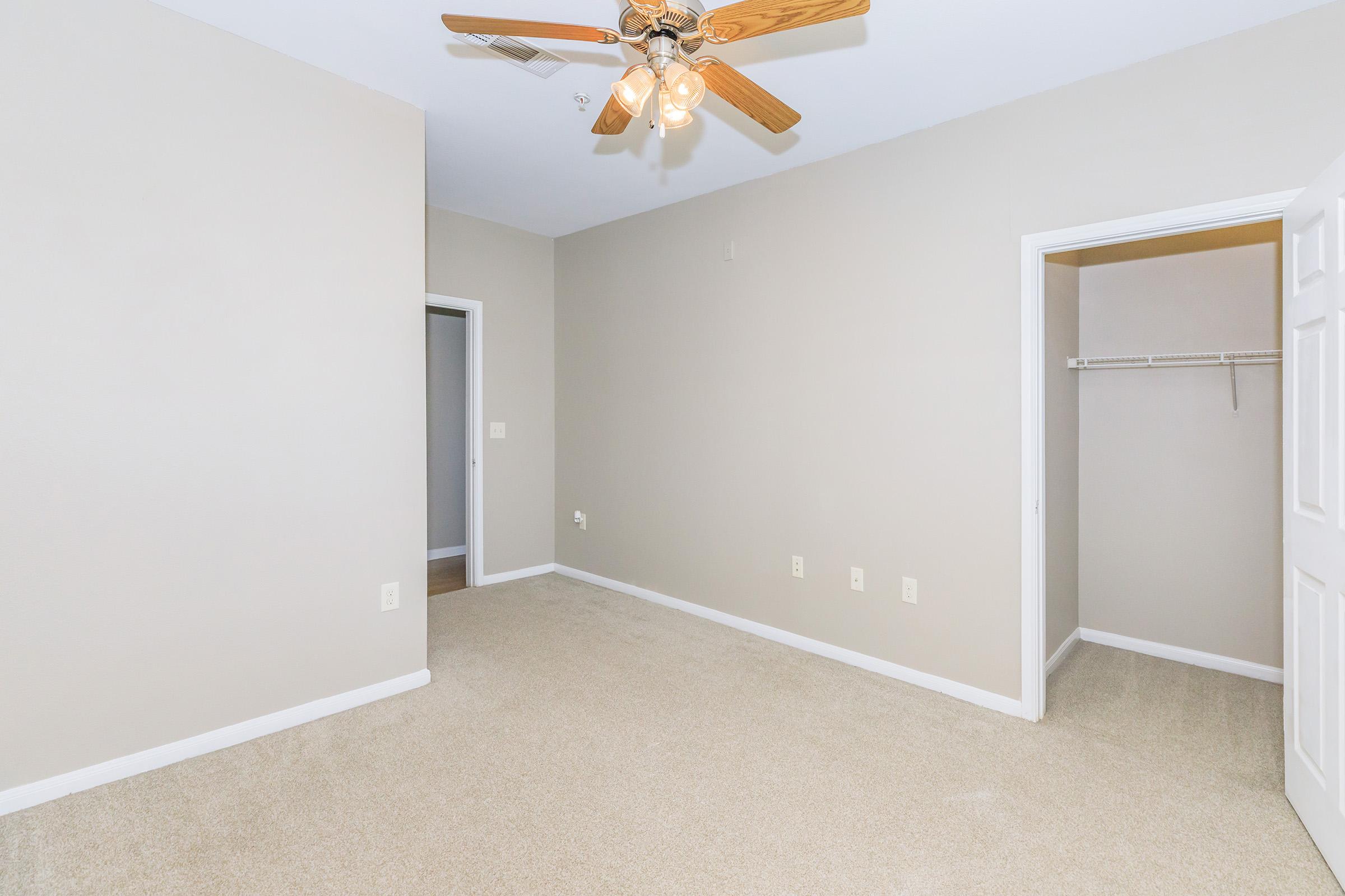 A spacious, unfurnished bedroom featuring light beige walls and carpeting. A ceiling fan with wooden blades is installed, providing ventilation. The room has a doorway leading to a closet on one side and another door leading to an adjacent room. The overall atmosphere is bright and inviting.