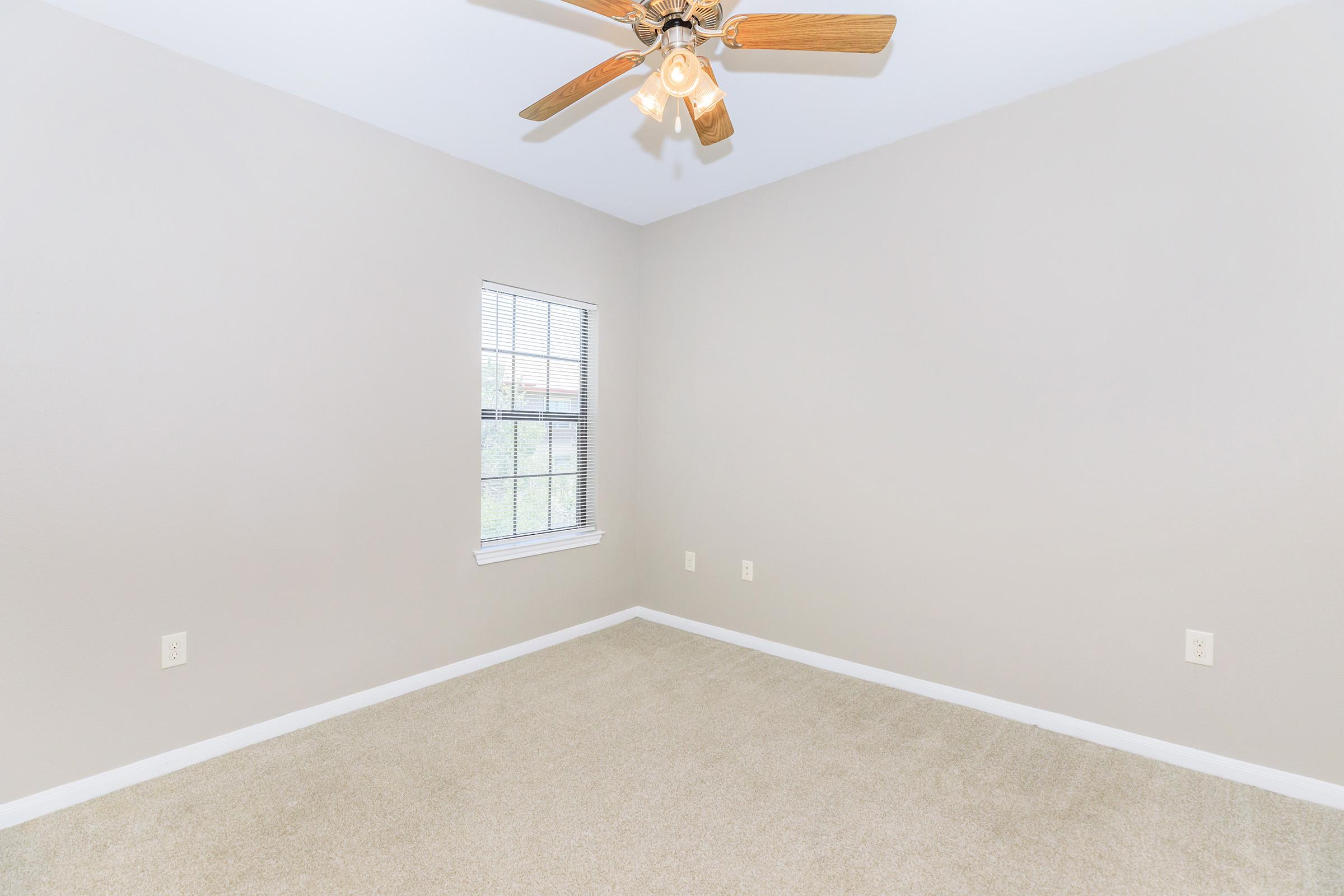 A small, empty bedroom featuring light beige walls and carpet. It has a ceiling fan with wooden blades and a window with blinds, allowing natural light to enter. The space appears clean and ready for furniture or decoration.