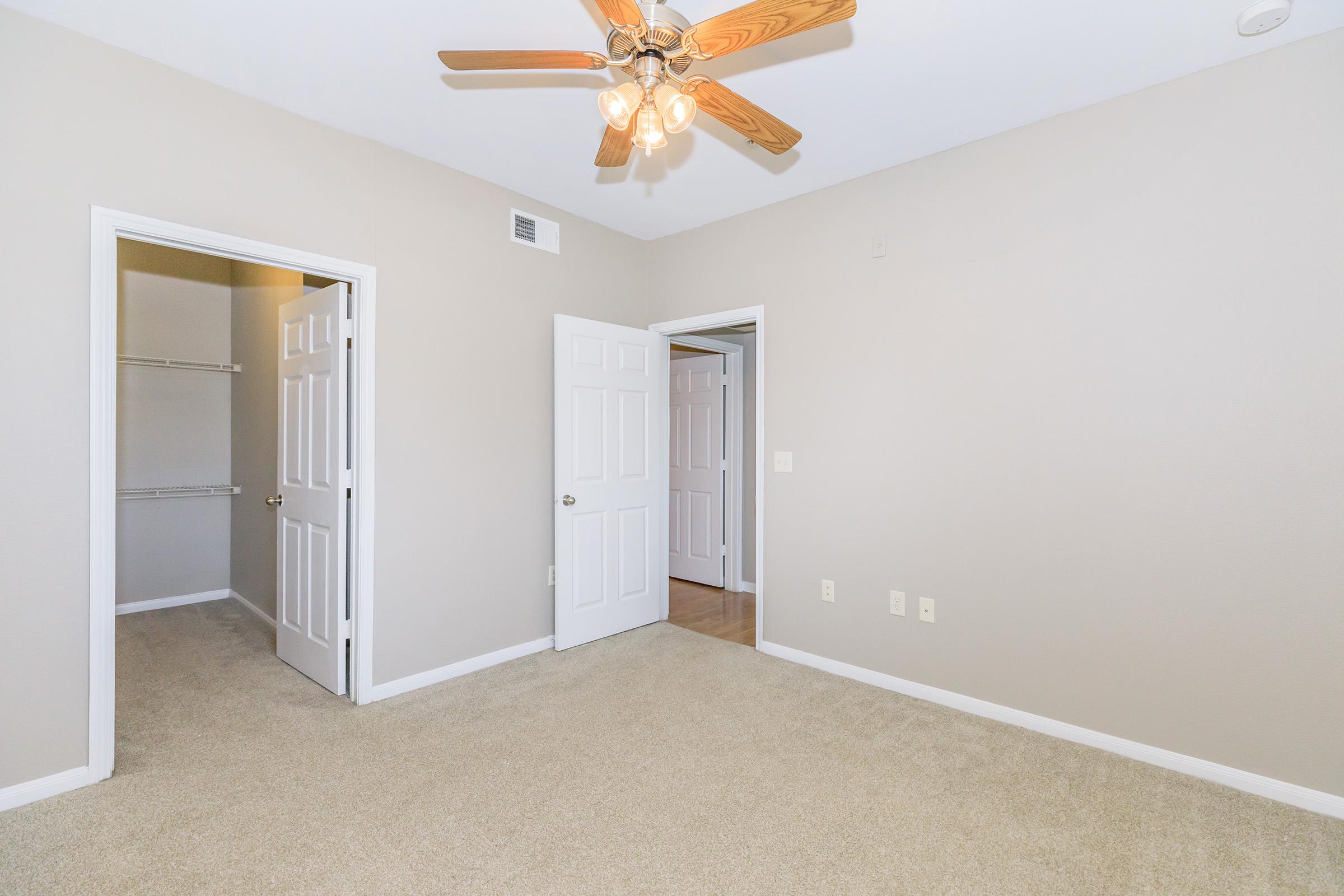 Empty room with light beige walls and carpet. There are two open doors leading to closets, and a ceiling fan with wooden blades is visible. The space is well-lit, showcasing a clean and simple layout suitable for various uses.