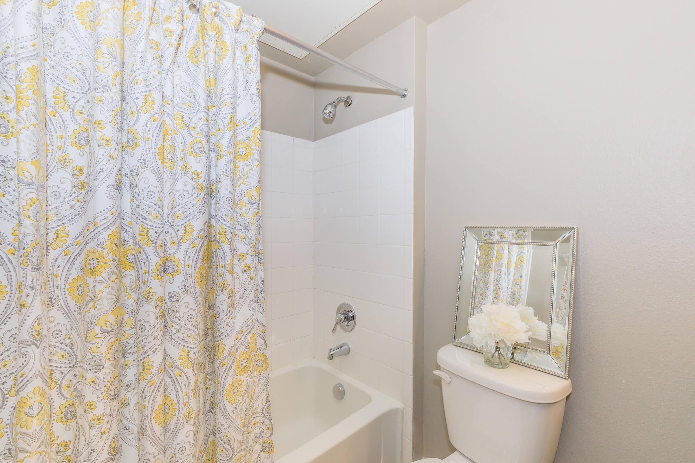 A clean bathroom featuring a white bathtub with a yellow and gray patterned shower curtain. There is a chrome showerhead, a white toilet beside the bathtub, and a decorative silver-framed mirror above a white floral arrangement. The walls are painted a light gray color.