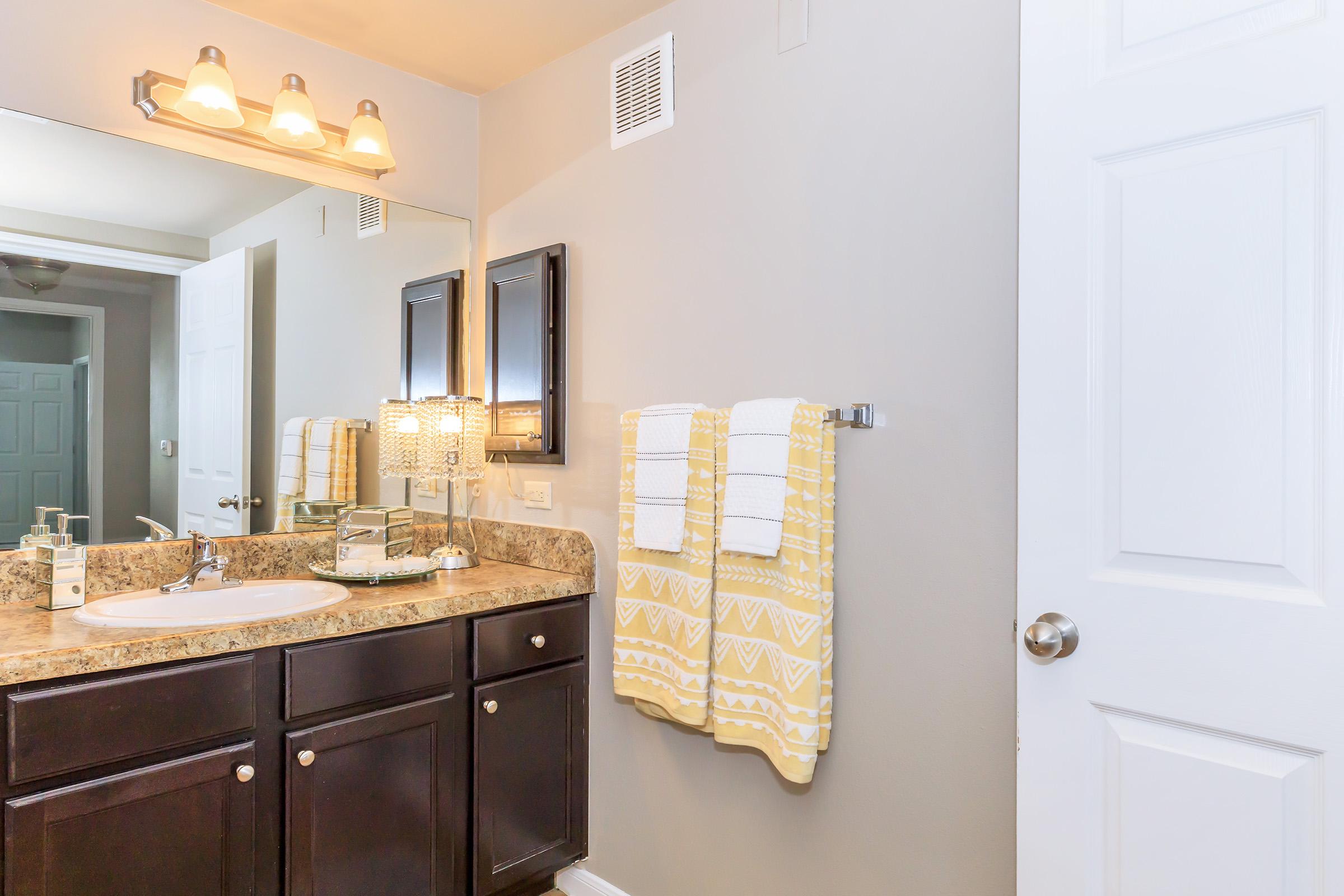 Bright and modern bathroom featuring a beige countertop with a sink, dark wooden cabinets, and a wall mirror with lights above. Two neatly hung yellow and white patterned towels are displayed. A clean white door leads out of the room, contributing to the overall light and airy atmosphere.