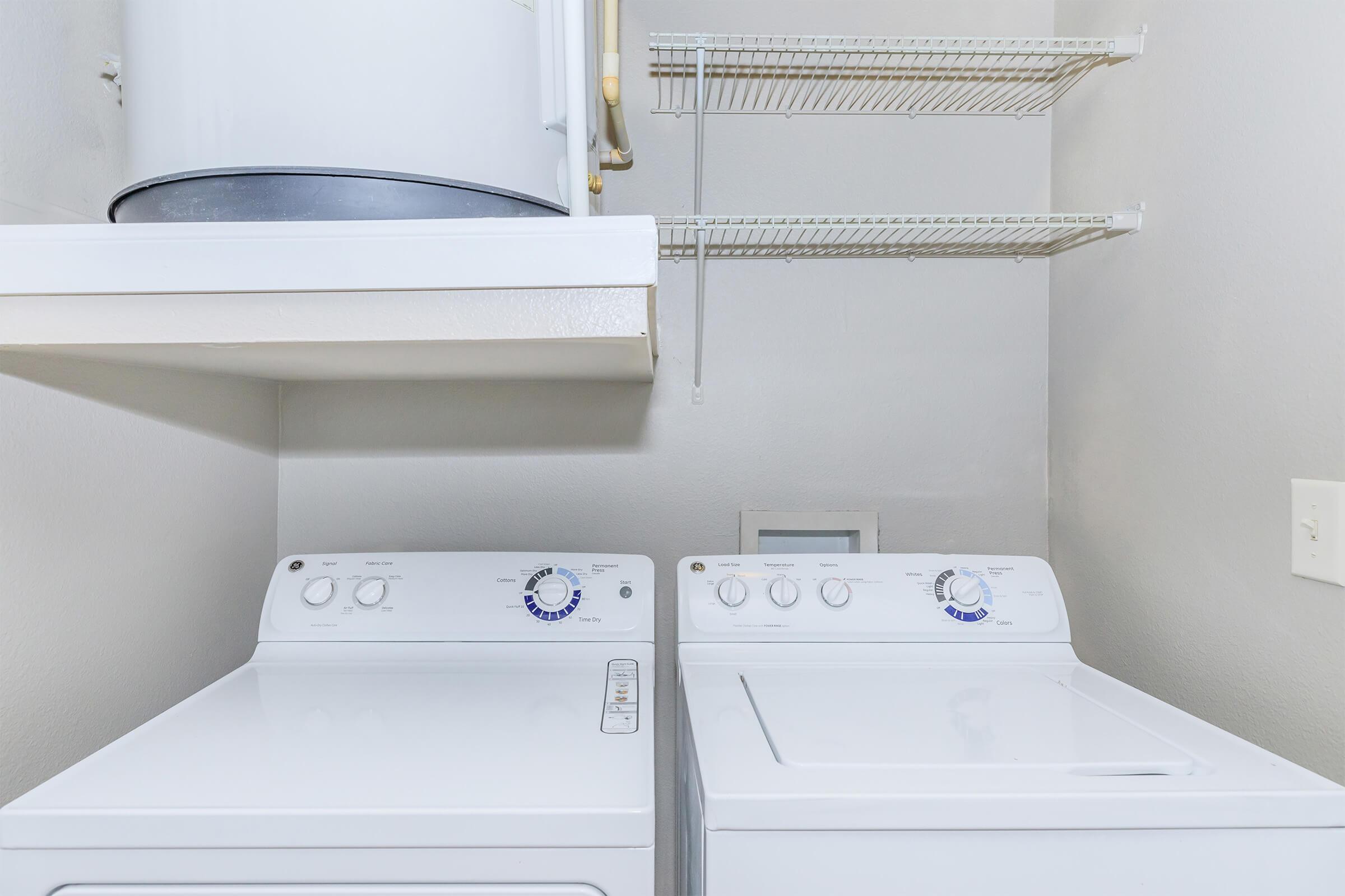 A clean laundry room featuring a white washer and dryer side by side. Above them, there is a shelf for storage. A white water heater is visible in the background, and the walls are painted a light gray. The overall space is tidy and organized, suitable for laundry tasks.