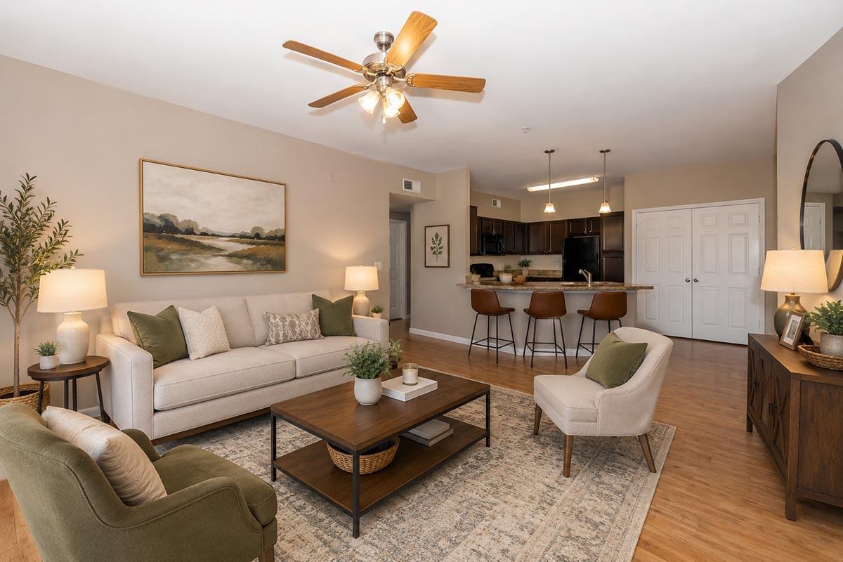 A cozy living room with a neutral color palette featuring a beige sofa, green and cream cushions, and a wooden coffee table. There’s a rug under the furniture, two lamps on side tables, and a painting above the sofa. In the background, a kitchen bar with three stools is visible.