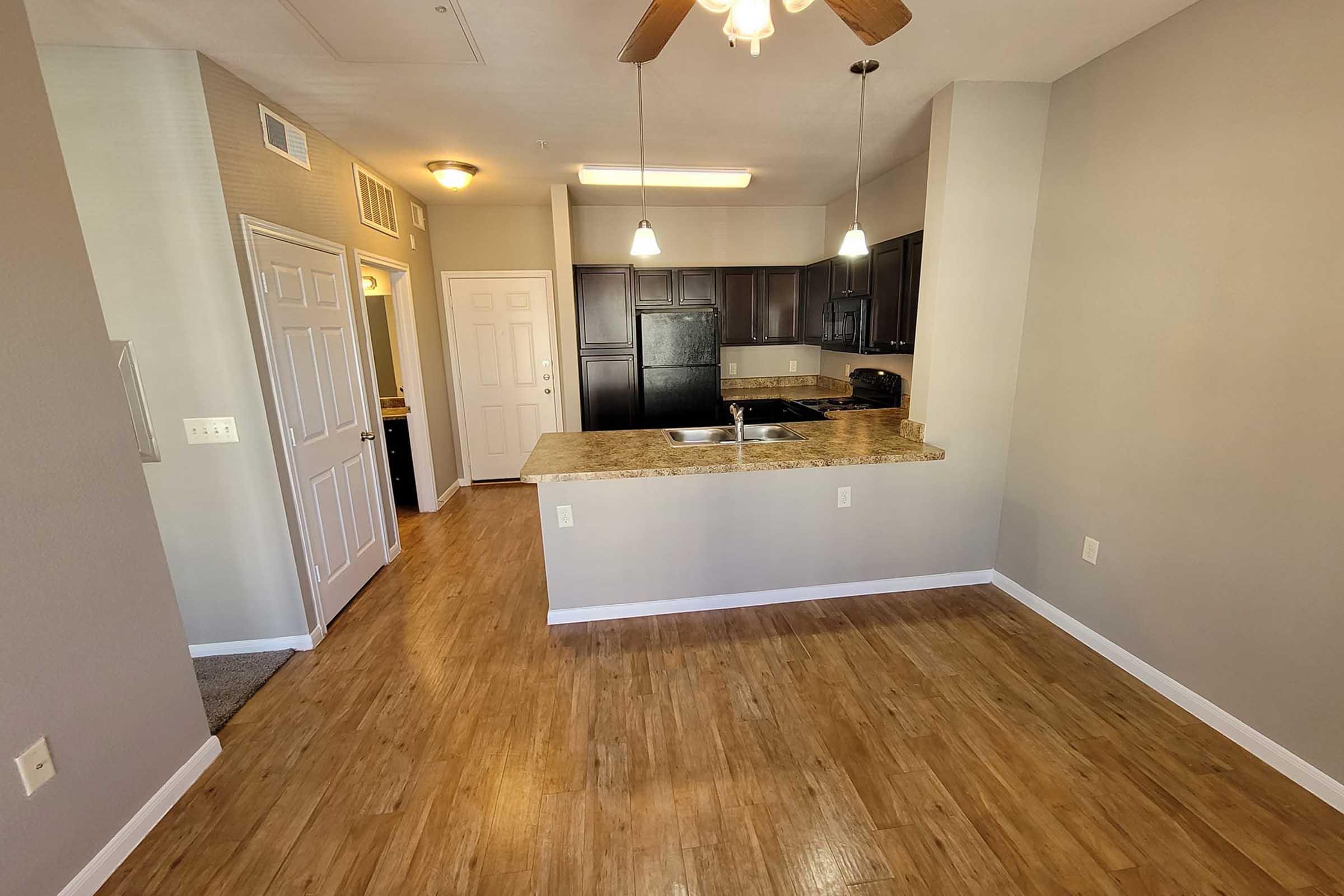 A spacious kitchen and living area featuring wood laminate flooring, a ceiling fan, and dark cabinetry. Countertops with a sink are visible, along with pendant lighting above. The layout includes doorways leading to other rooms.