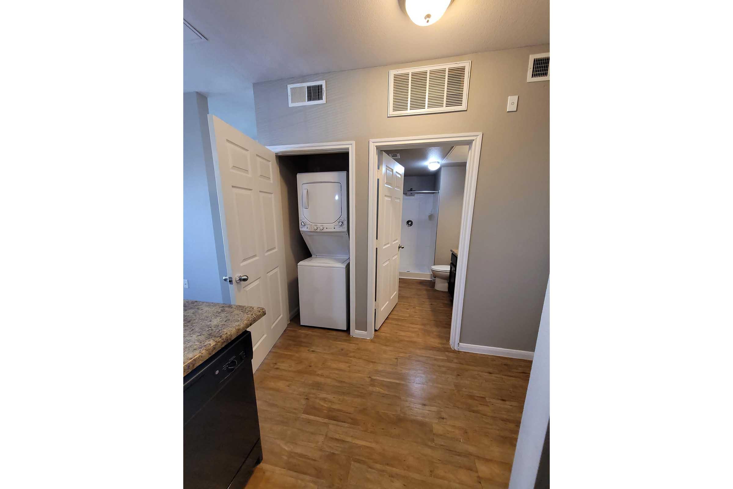 View of a hallway in an apartment featuring two closed doors; one door leads to a stackable washer and dryer, while the other opens to a bathroom. The space has light wooden flooring and neutral-colored walls.