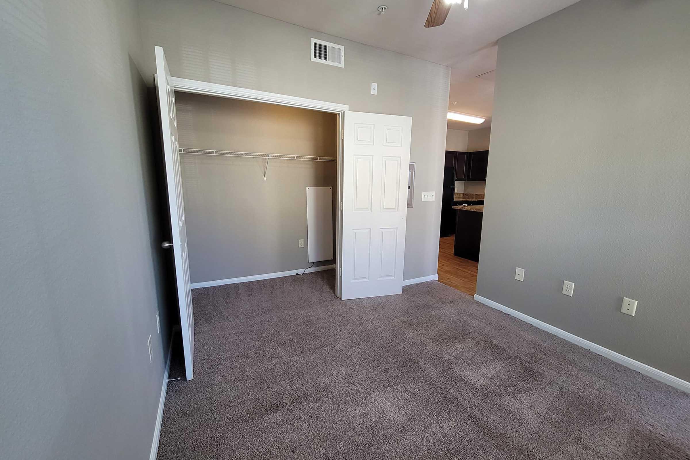 A vacant room with gray walls and carpet, featuring an open closet with shelving and a white door leading to another area. There is a ceiling fan above, and the background shows a blurred view of a kitchen or living area.