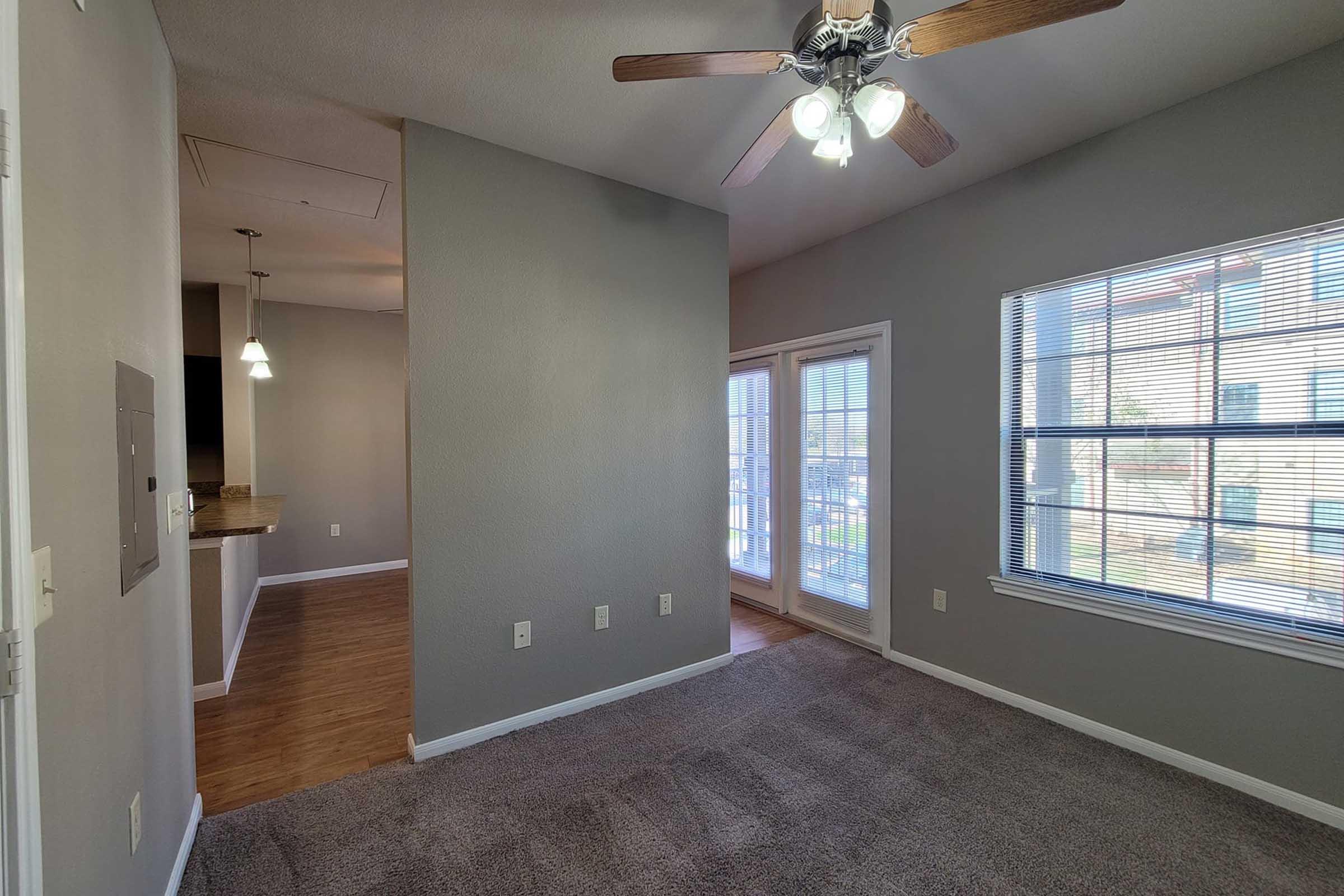 Interior view of a living space featuring gray walls, carpeted flooring, a ceiling fan with lights, and windows providing natural light. The layout includes a transition to a kitchen area visible in the background, and a sliding door leading to an outdoor space.