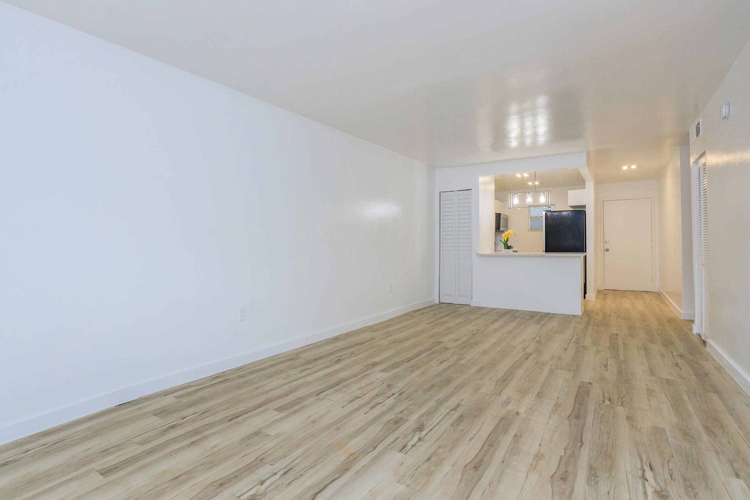 Spacious, empty room with light-colored wood flooring and white walls. The area features an open layout leading to a kitchen with modern appliances in the background. Natural light fills the space, enhancing the clean, minimalist aesthetic.