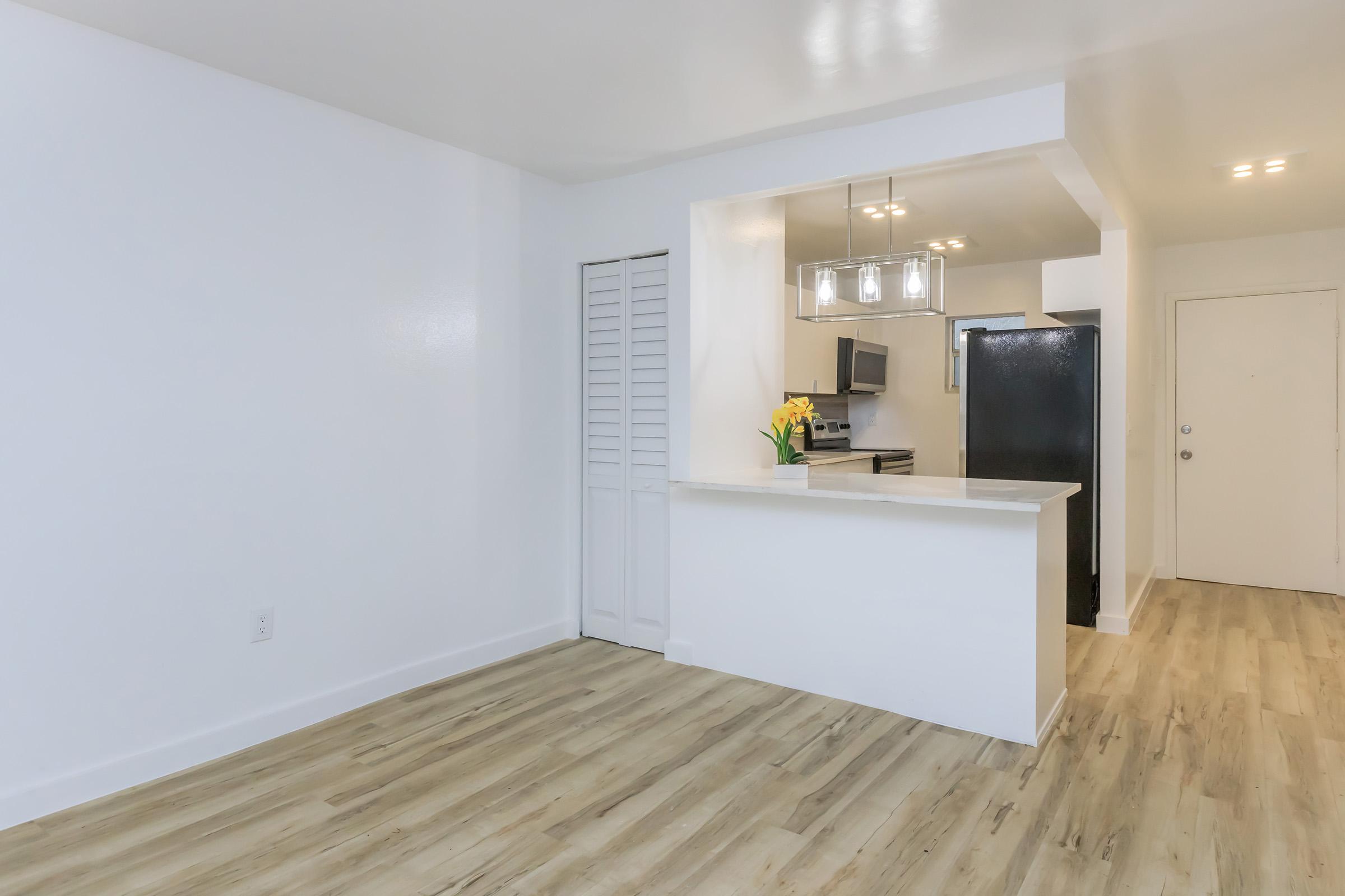 A modern, bright interior of a kitchen and living space featuring light-colored walls, a bar counter with flowers, and a black refrigerator. There are wooden floors, recessed lighting, and an open layout connecting the kitchen to the living area. A door leads to another room or area.