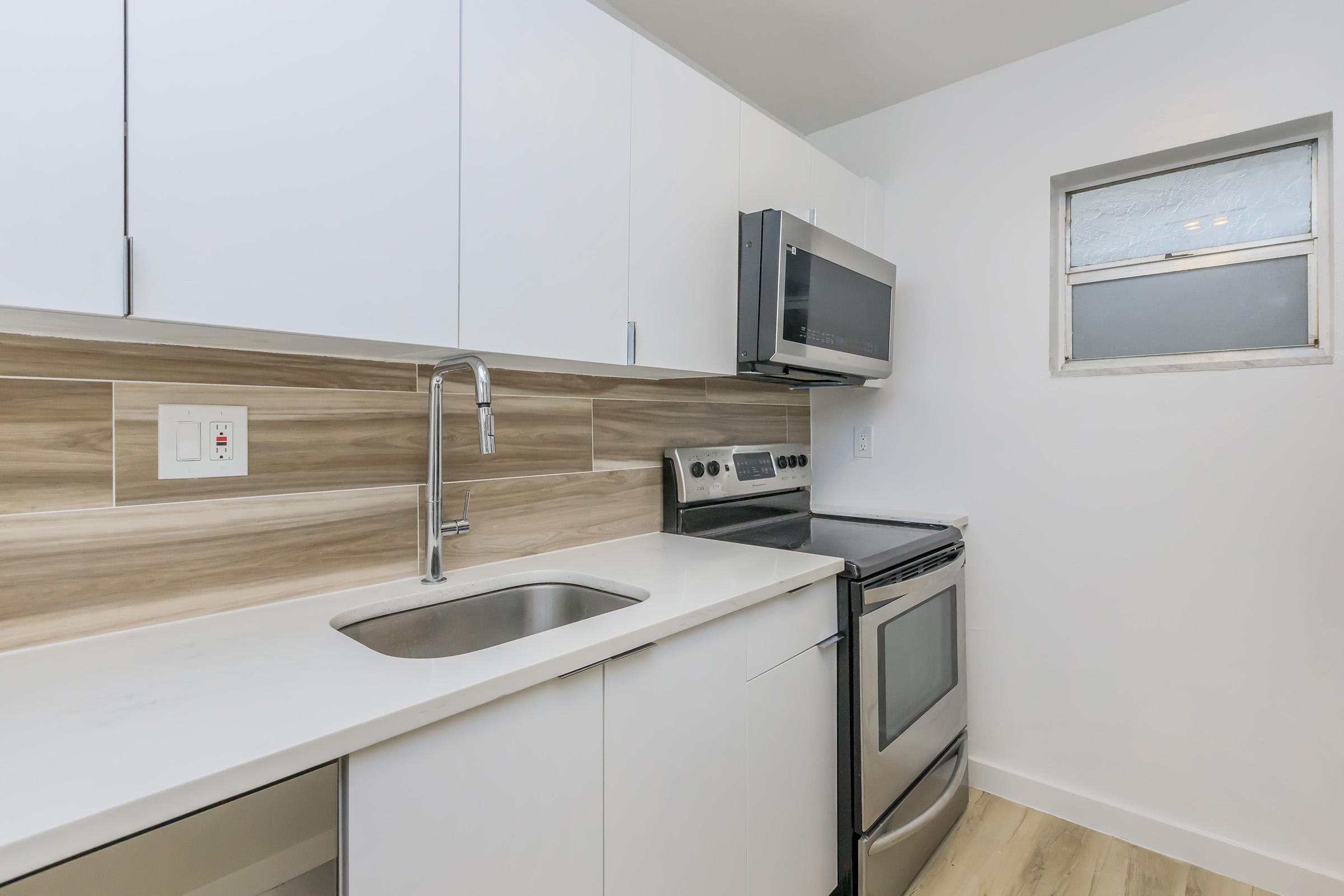 A modern kitchen featuring white cabinets, a stainless steel microwave, and an oven. The countertop is a light color with a sleek design, and there's a single basin sink. A small window above the sink allows natural light, while the walls are painted in a clean, neutral tone.