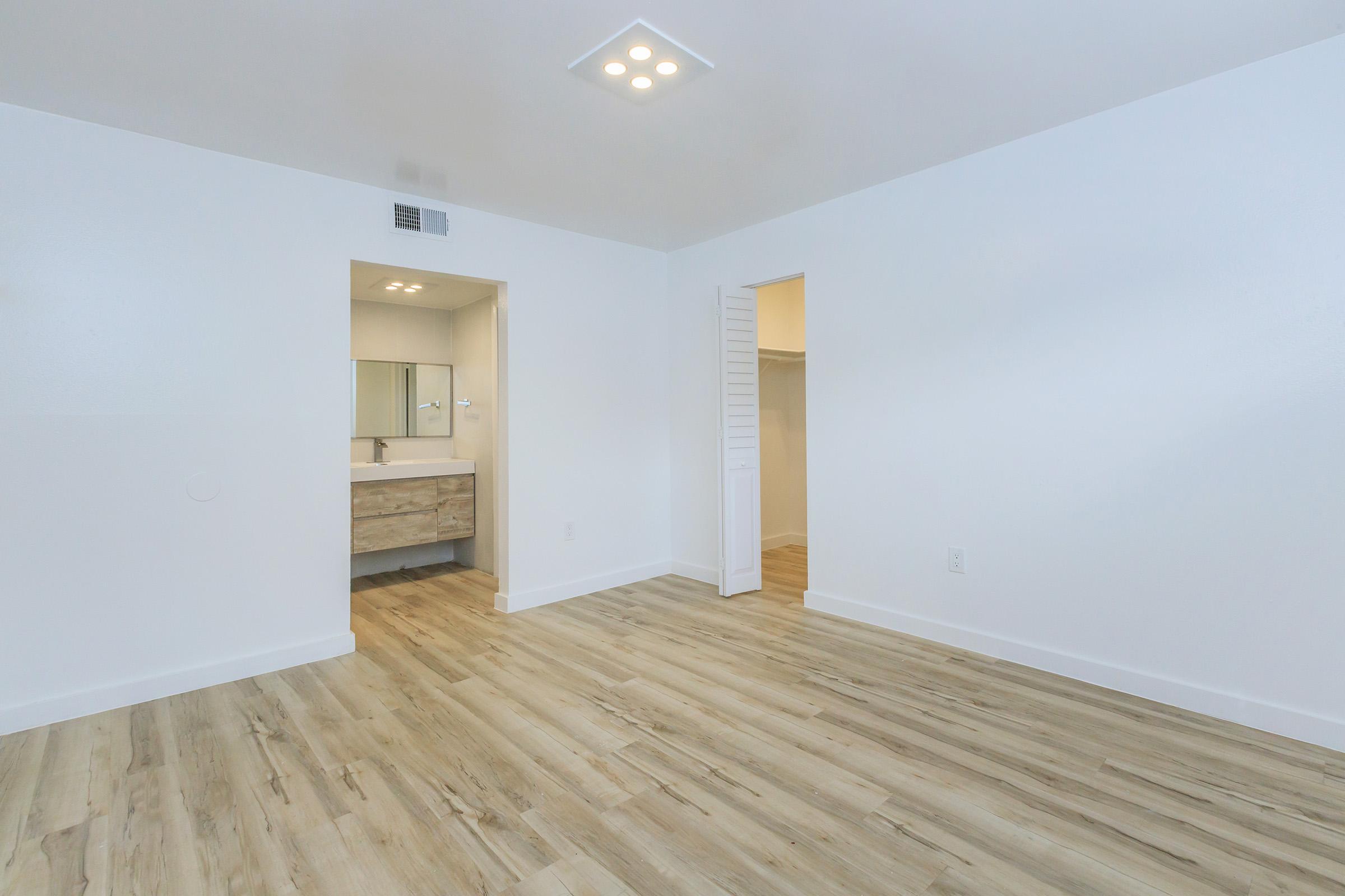 A spacious, modern room with light wood flooring. The walls are white, and there's a doorway leading to a bathroom with a vanity and mirror. An open closet is visible in the corner, providing additional storage space. The ceiling features recessed lighting for a bright atmosphere.