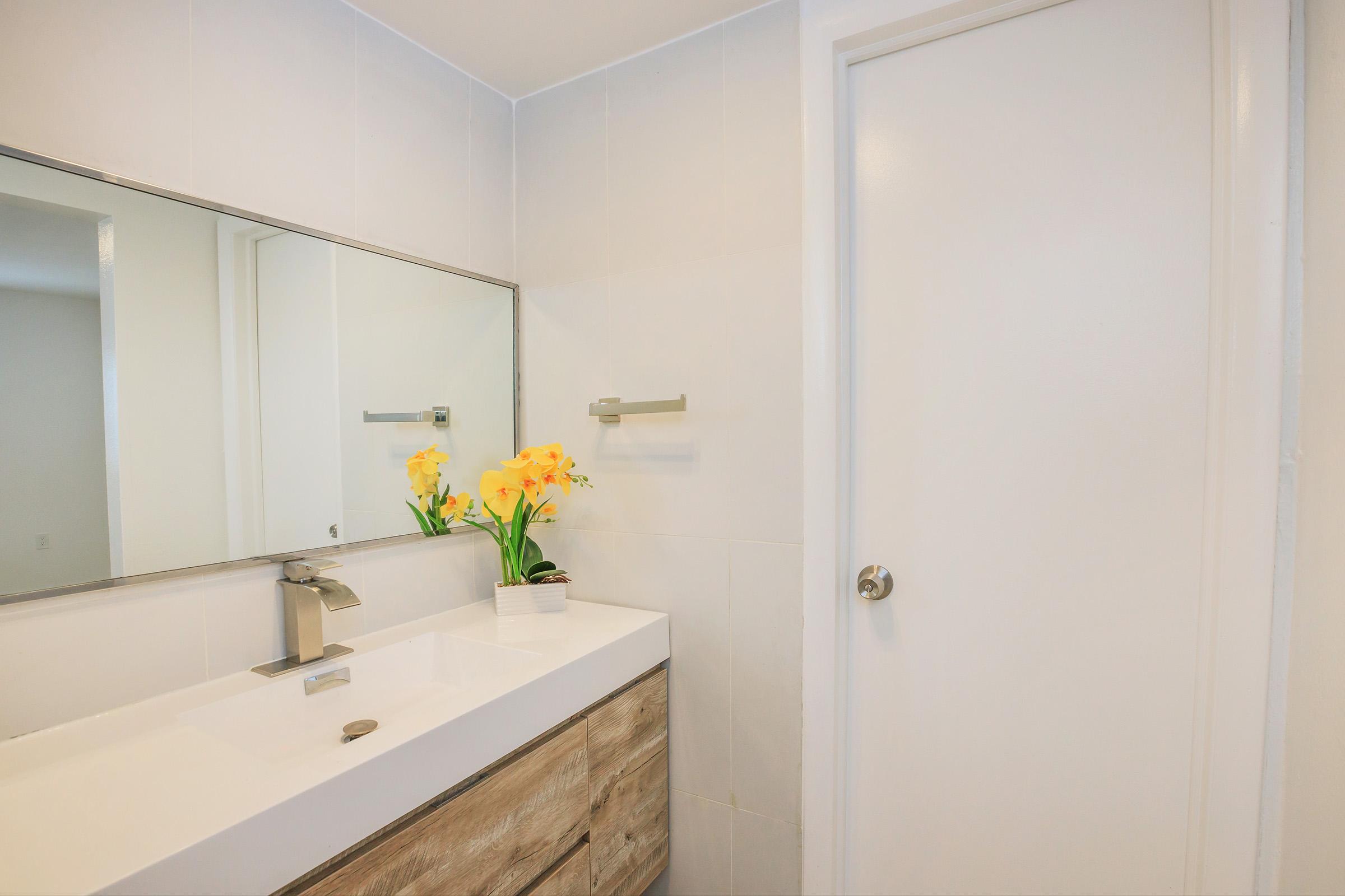 A modern bathroom featuring a white sink and wooden vanity below a large mirror. Bright yellow flowers in a small pot sit on the vanity. A clean, white door is visible on the right, and a light-colored wall completes the simple, fresh design.