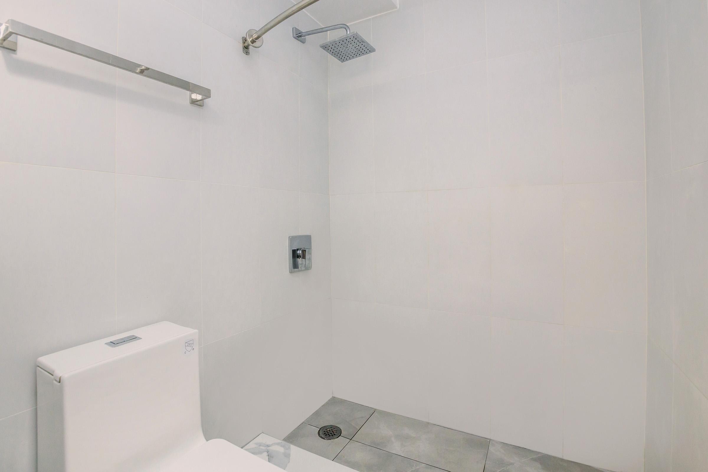 A minimalistic bathroom featuring a gray tiled floor, a white toilet, a stainless steel grab bar, and a modern shower head mounted on a white tiled wall. The space has a clean, contemporary design with an abundance of white surfaces and natural light.