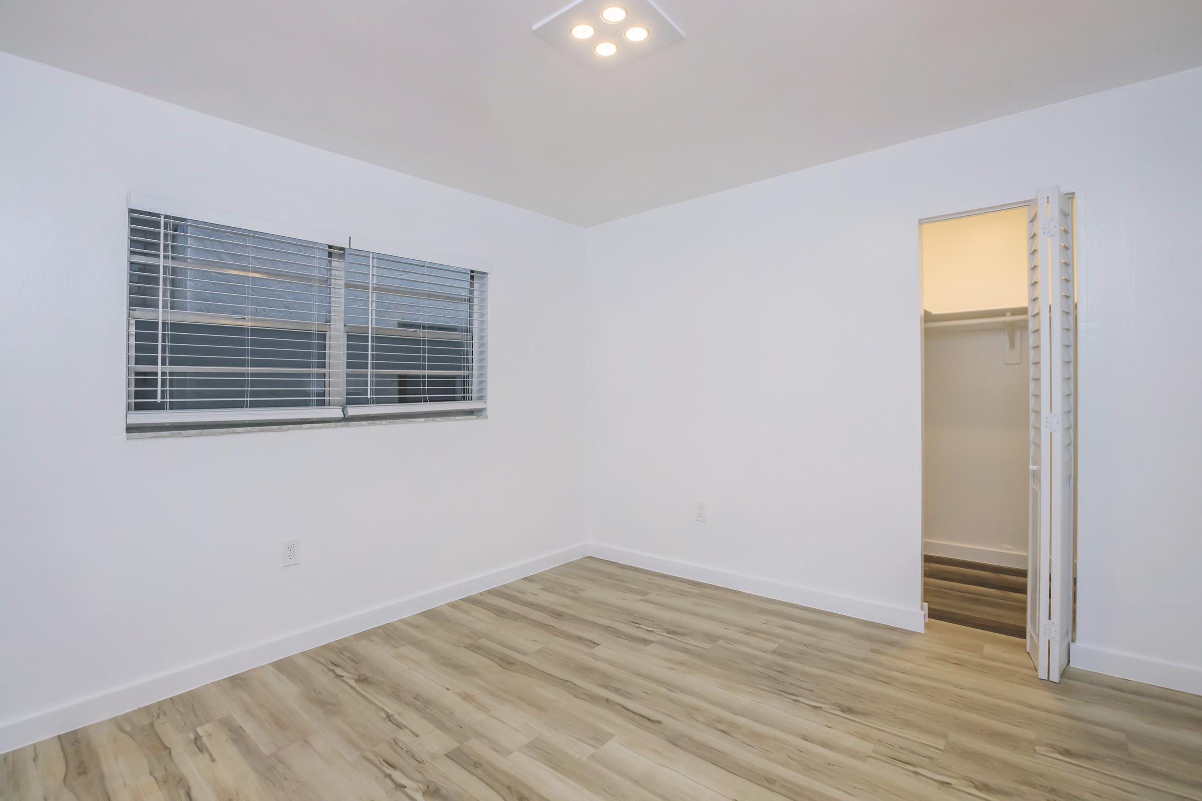 A minimalistic room with white walls and a light-colored wooden floor. It features a window with blinds on one wall and an open closet doorway on the right. The ceiling has recessed lighting, creating a bright and airy atmosphere.