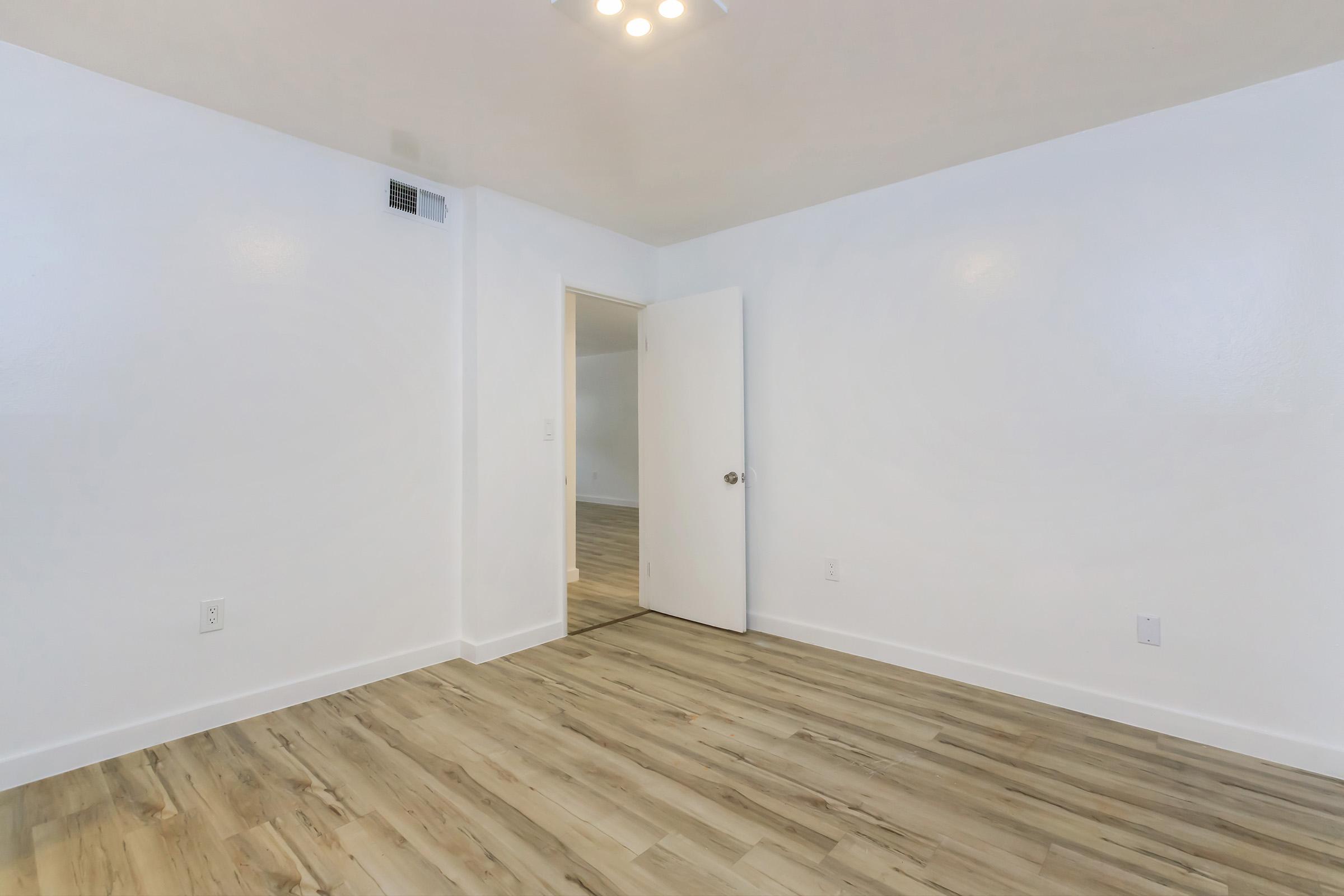 A spacious, empty room with light-colored walls, wooden flooring, and a door leading to another room. The ceiling has simple lighting fixtures, and the overall atmosphere is bright and clean, creating a blank canvas for interior design.