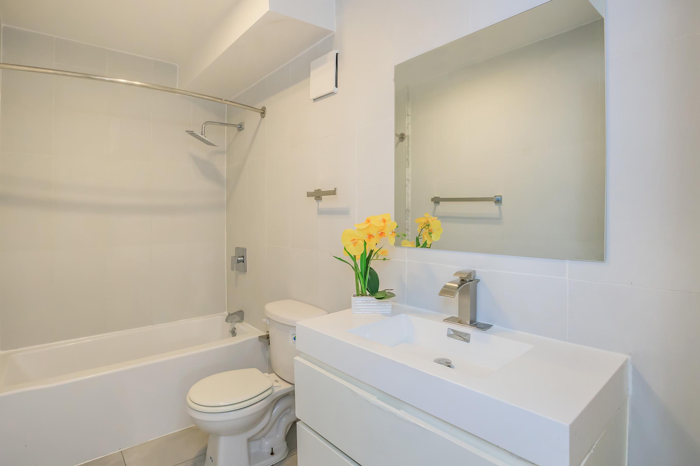 A modern bathroom featuring a white bathtub, a toilet, a sleek white vanity with a sink, and a large mirror. Yellow flowers in a small vase add a touch of color to the neutral-toned walls and fixtures, creating a bright and clean ambiance.