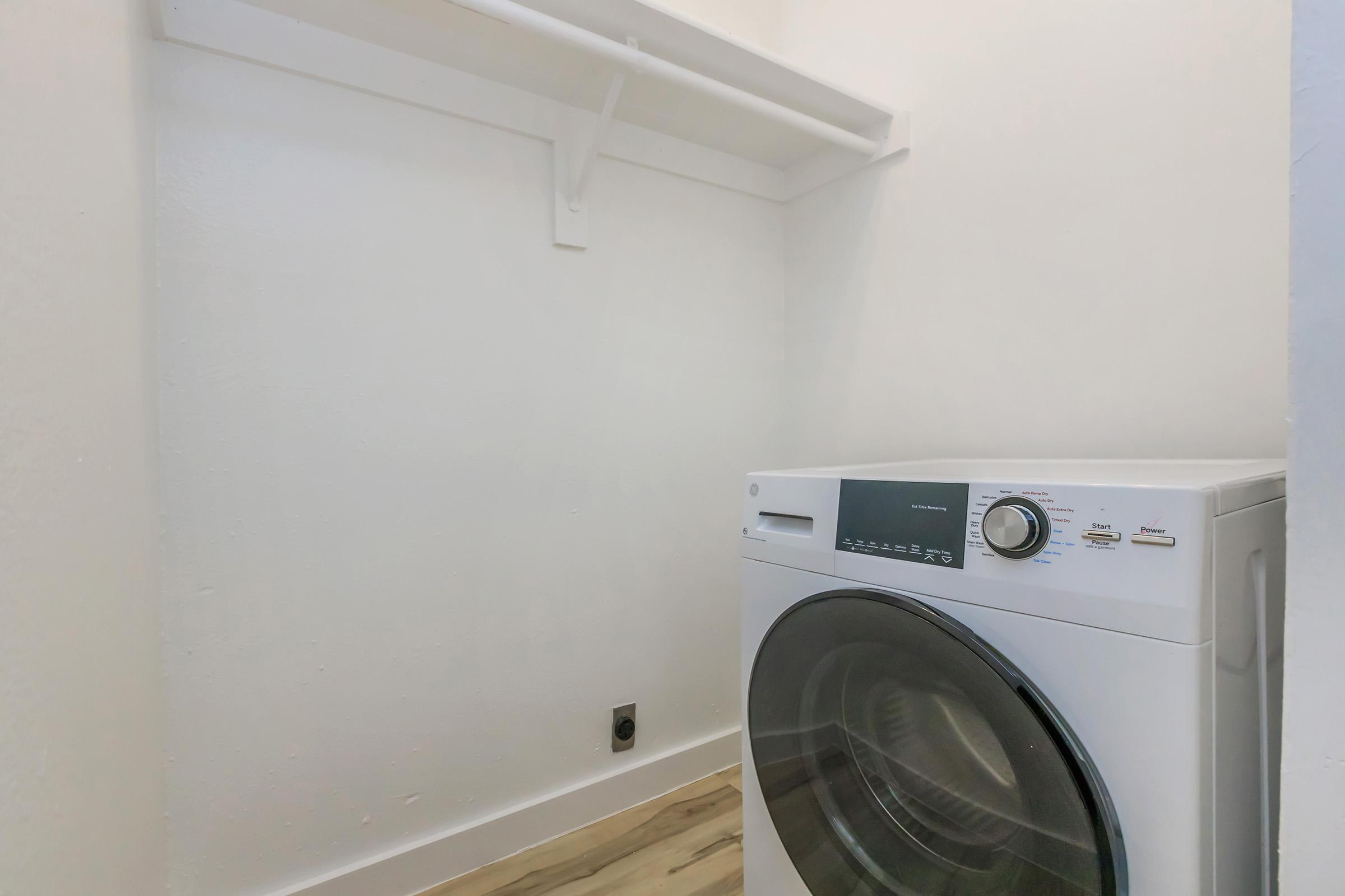 A clean laundry room featuring a modern washing machine against a white wall, with a shelf above it for storage. The floor has a light wood finish, adding a warm touch to the space.