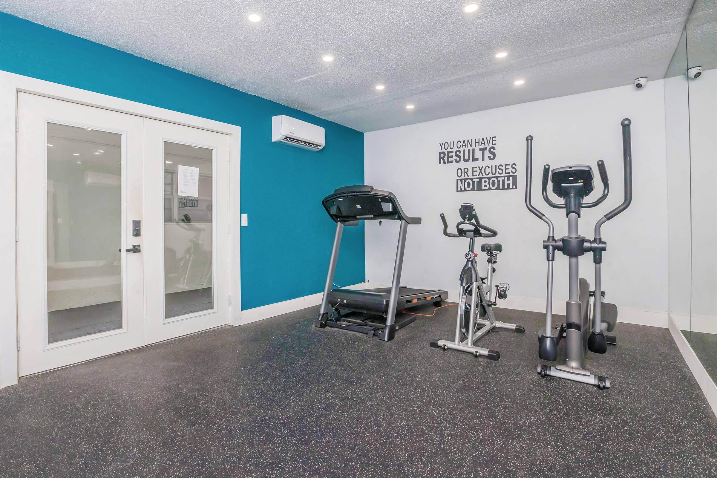 A modern home gym featuring a treadmill and an elliptical machine positioned against a blue accent wall. The room is well-lit with recessed lighting, and there is motivational wall art that reads, "You can have results or excuses, not both." Mirrored walls enhance the sense of space.