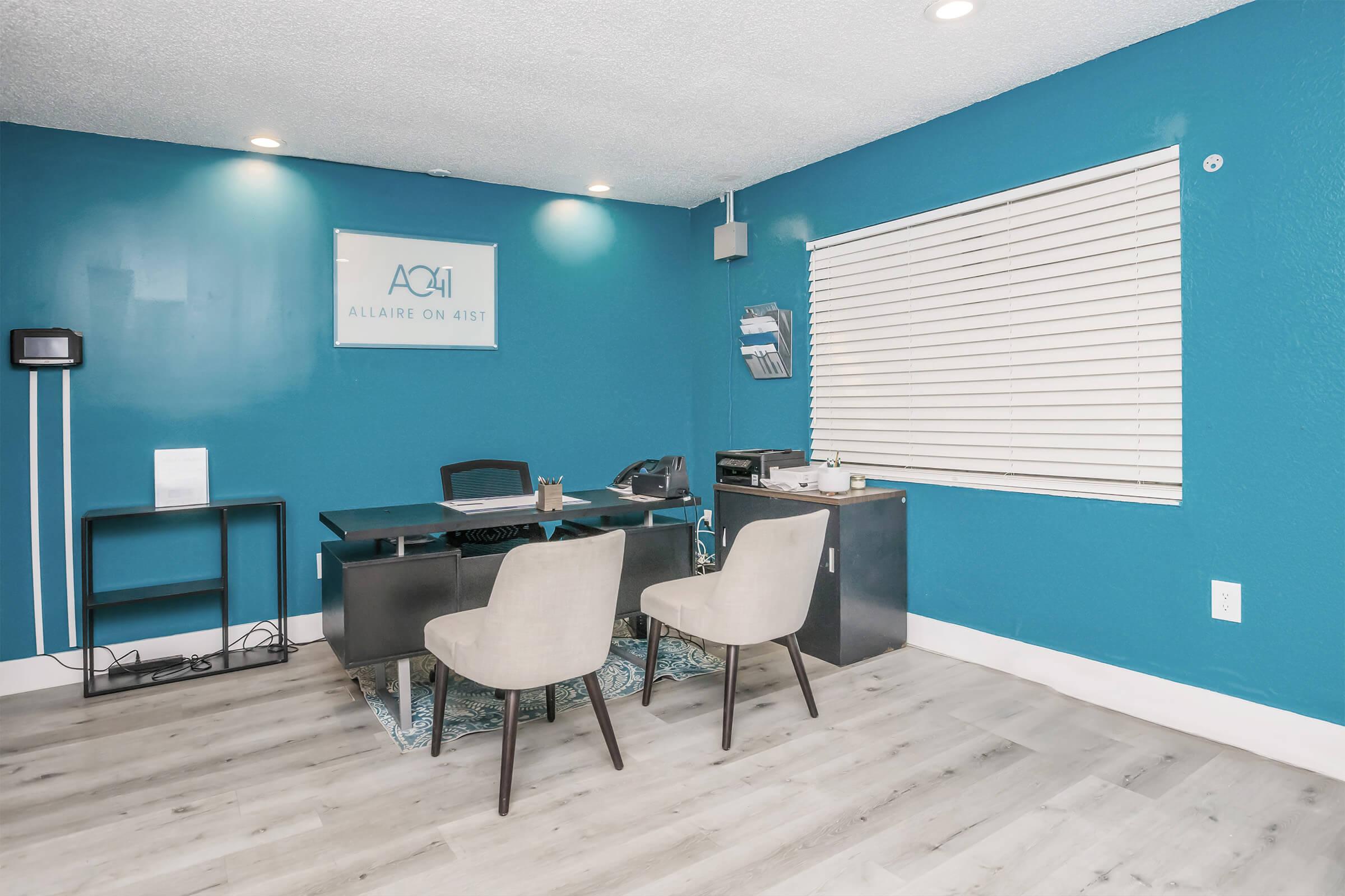 A modern office space featuring a teal wall, a desk with a chair, and two light-colored chairs. There's a window with blinds allowing natural light, and a logo on the wall. The floor is light-colored wood, adding to the contemporary design.
