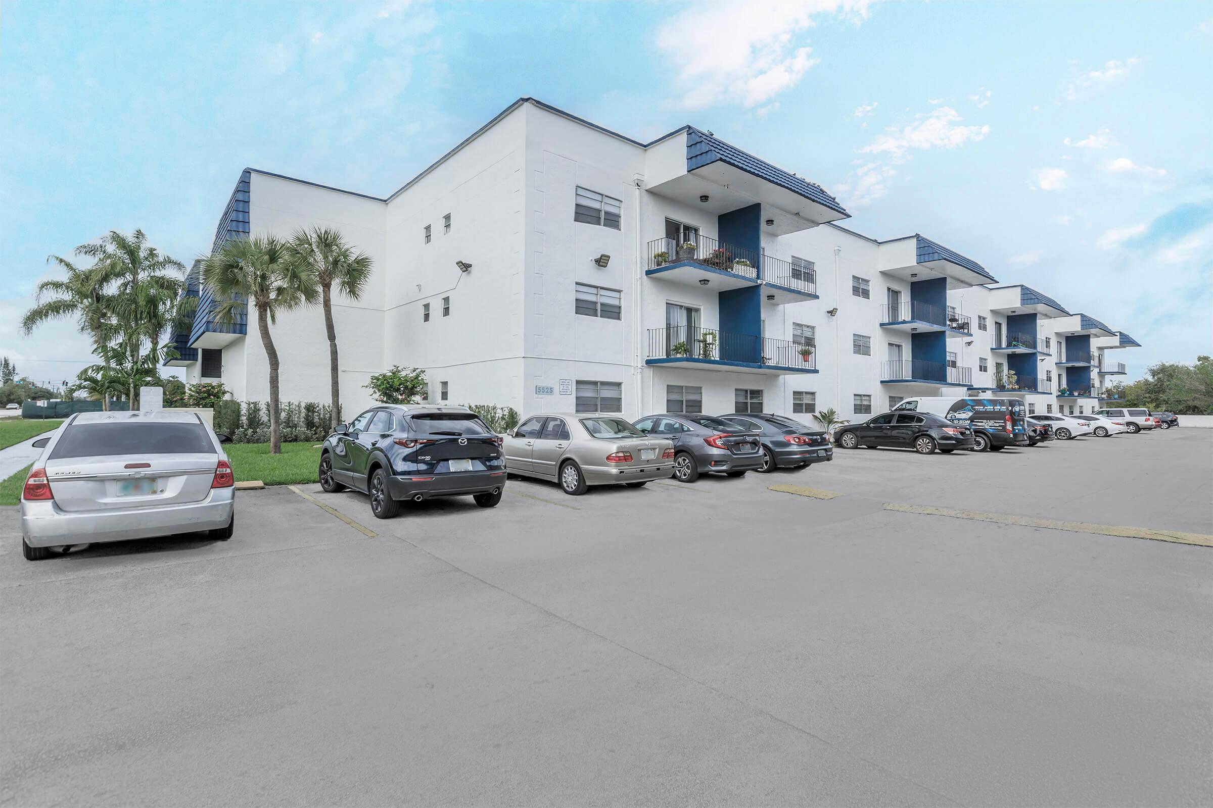 A modern apartment building with a white exterior and blue accents, featuring multiple balconies. Several cars are parked in front of the building, with neatly landscaped palm trees in the vicinity and a clear blue sky above.