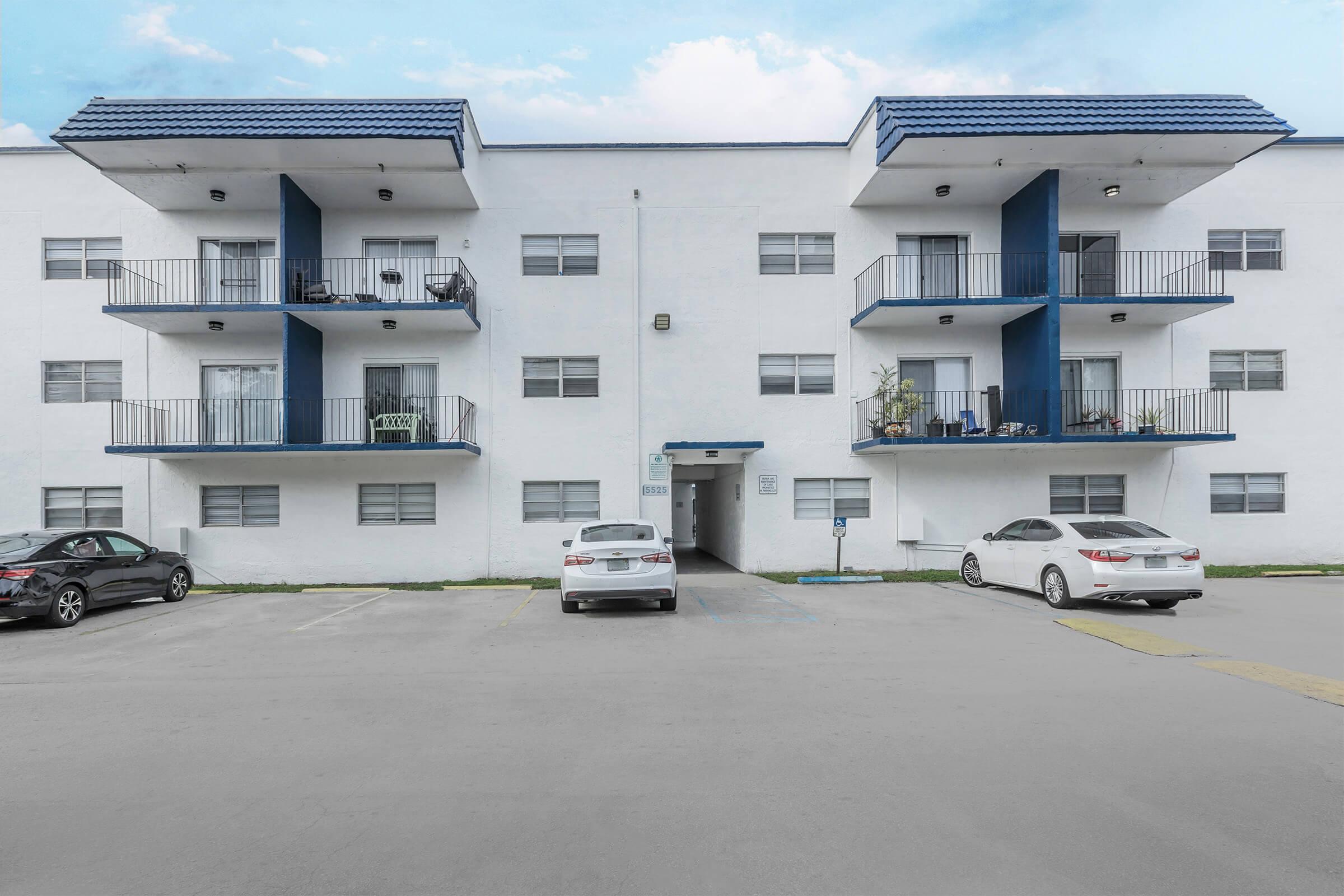 A three-story apartment building with a white exterior, blue accents, and balconies. The front features parked cars in a lot, with an entrance visible. Some balconies are decorated with plants. The sky is partly cloudy, adding to the overall residential setting.