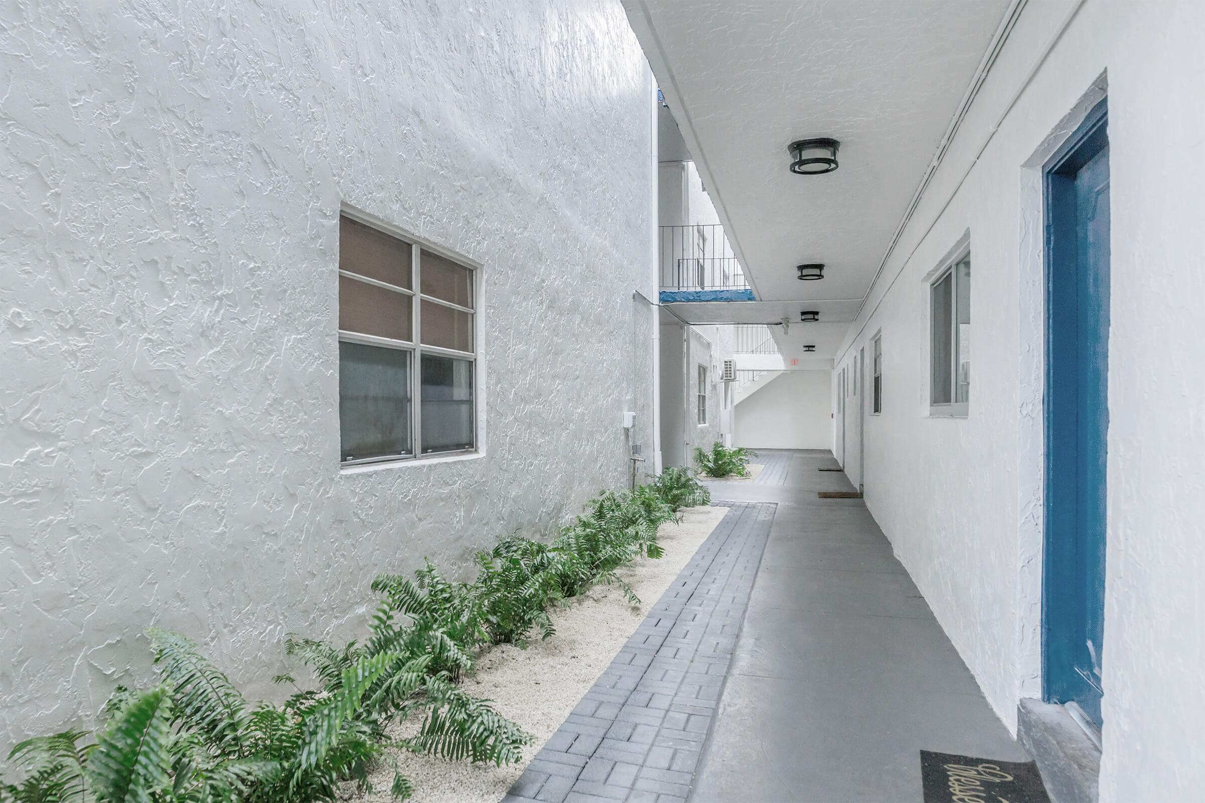 Narrow corridor between white textured walls, featuring windows on one side and a blue door at the far end. The ground is paved with gray tiles and adorned with small patches of greenery and white pebbles, creating a clean and inviting atmosphere.