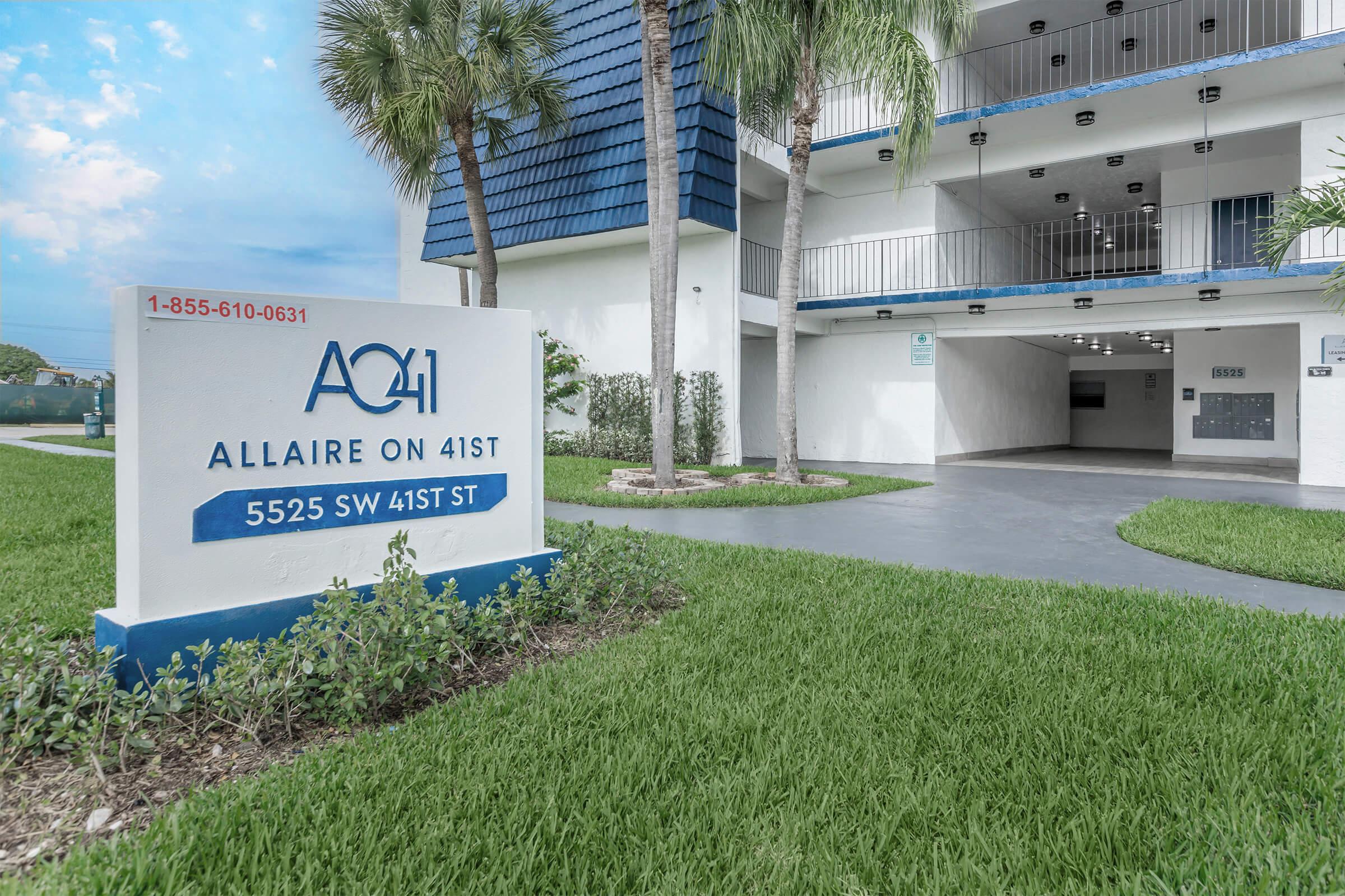 Sign for "Allaire on 41st" with the address "5525 SW 41st St" displayed prominently in a well-maintained outdoor space featuring green grass and palm trees. The building has a modern architectural design, with balconies visible on the upper levels and a blue roof.