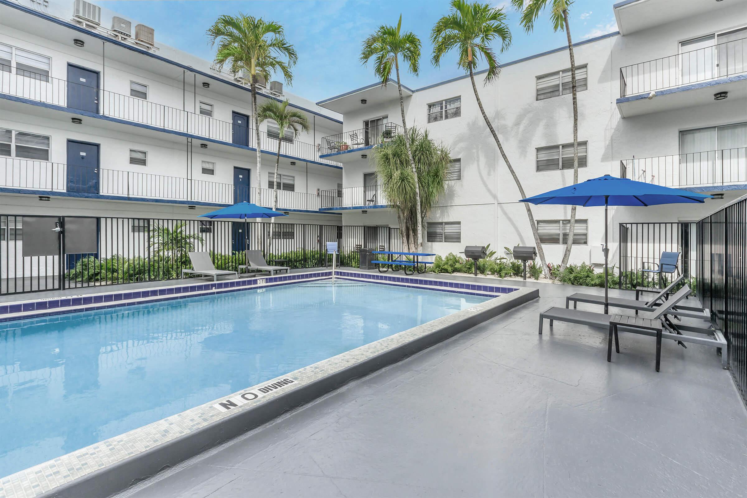 A view of a modern apartment complex featuring a swimming pool in the center. Surrounding the pool are lounge chairs and blue umbrellas. Palm trees provide a tropical ambiance, and the buildings in the background are white with blue accents. The area appears inviting and well-maintained.