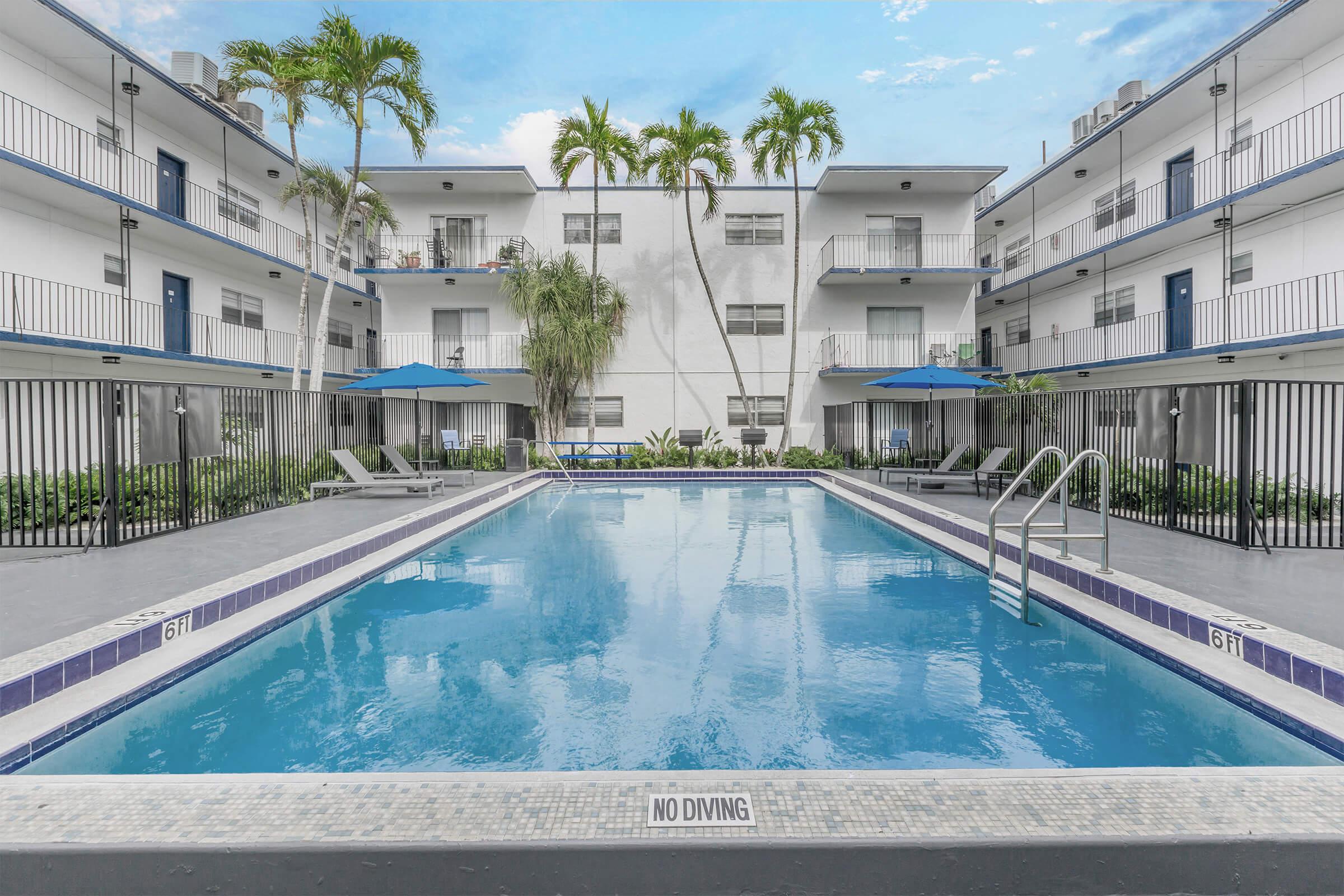 A modern hotel courtyard featuring a clear blue swimming pool surrounded by lounge chairs and umbrellas. The pool area is enclosed with a black fence, and the hotel buildings have balconies. Lush greenery and palm trees add to the tropical ambiance, with a bright blue sky overhead.