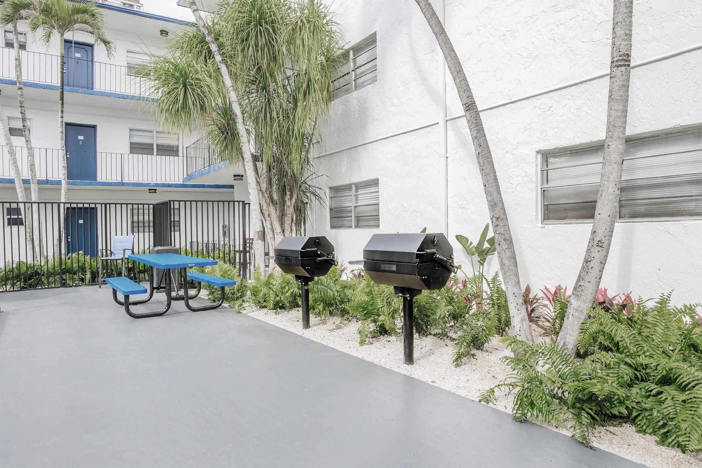 Outdoor area featuring two black charcoal grills on stands, surrounded by lush greenery and palms. There is a blue picnic table nearby. The setting is part of a courtyard next to a white building with balconies. The atmosphere is inviting and suitable for barbecues or gatherings.