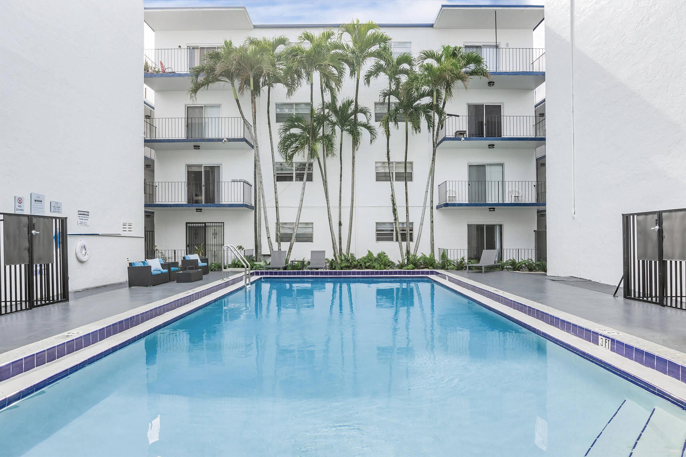 A clear swimming pool surrounded by palm trees and white buildings with balconies. The pool area features lounge chairs and has a clean, inviting appearance, ideal for relaxation and leisure. The setting is bright and sunny, enhancing the tranquil atmosphere.