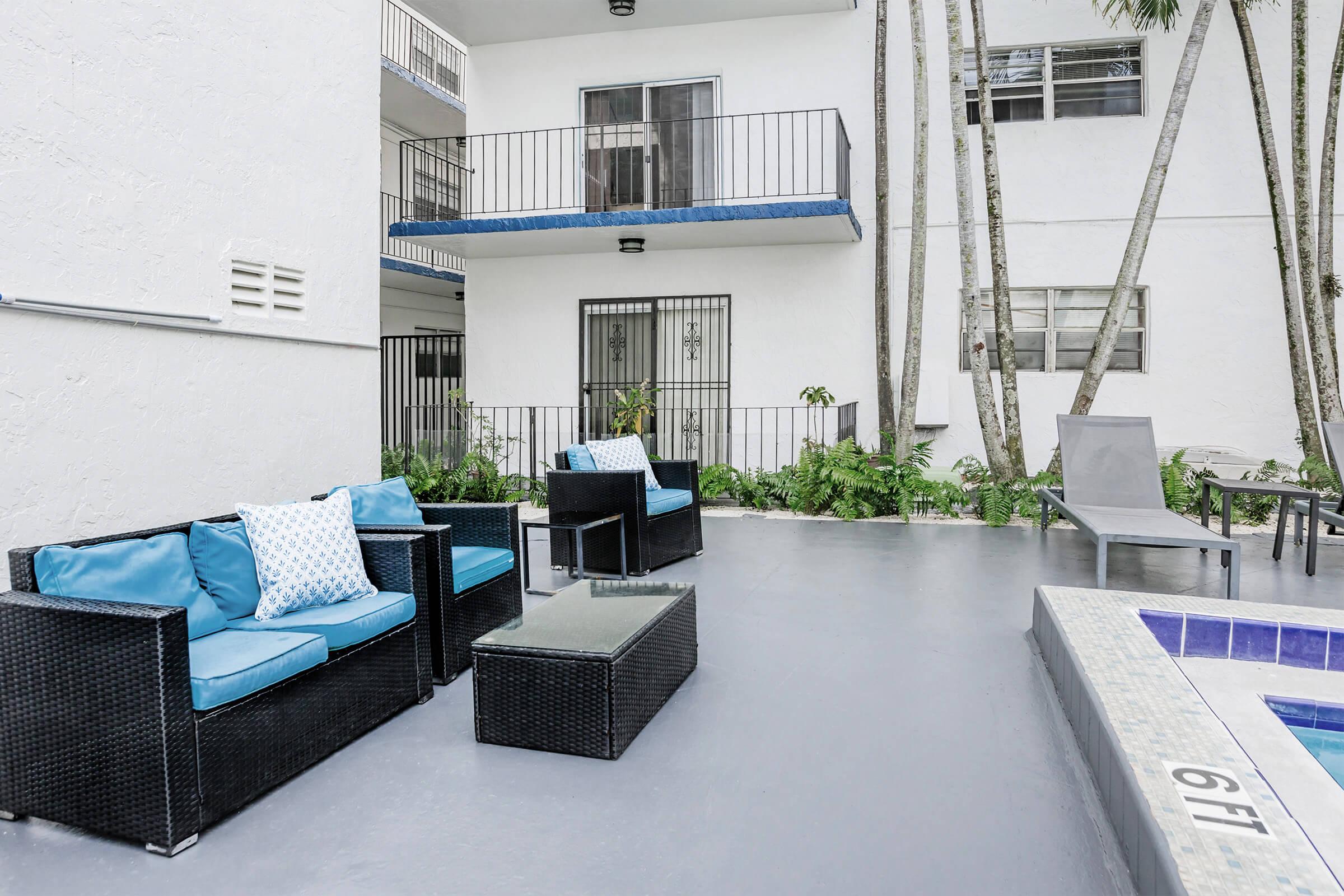 A modern outdoor space featuring a small seating area with blue cushions on wicker chairs, a glass-topped table, and a small hot tub nearby. The background includes a white building with balconies, greenery, and a concrete floor.