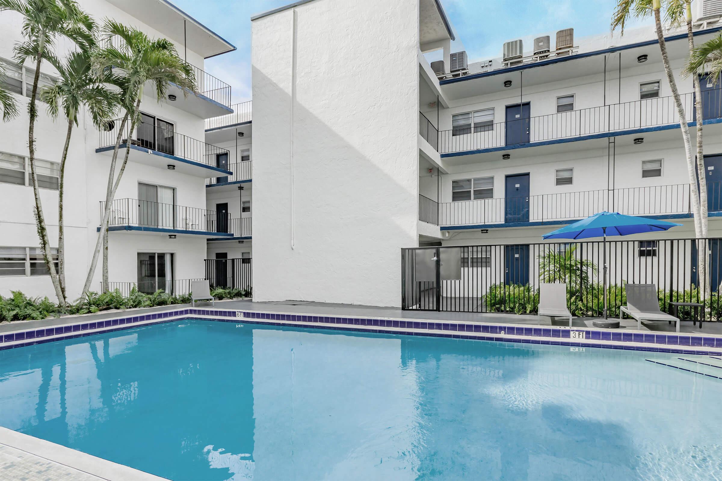 A clear blue swimming pool surrounded by an outdoor area featuring palm trees and lounge chairs. The pool area is enclosed by modern apartment buildings with balconies, and a large umbrella provides shade. The scene is welcoming and ideal for relaxation.