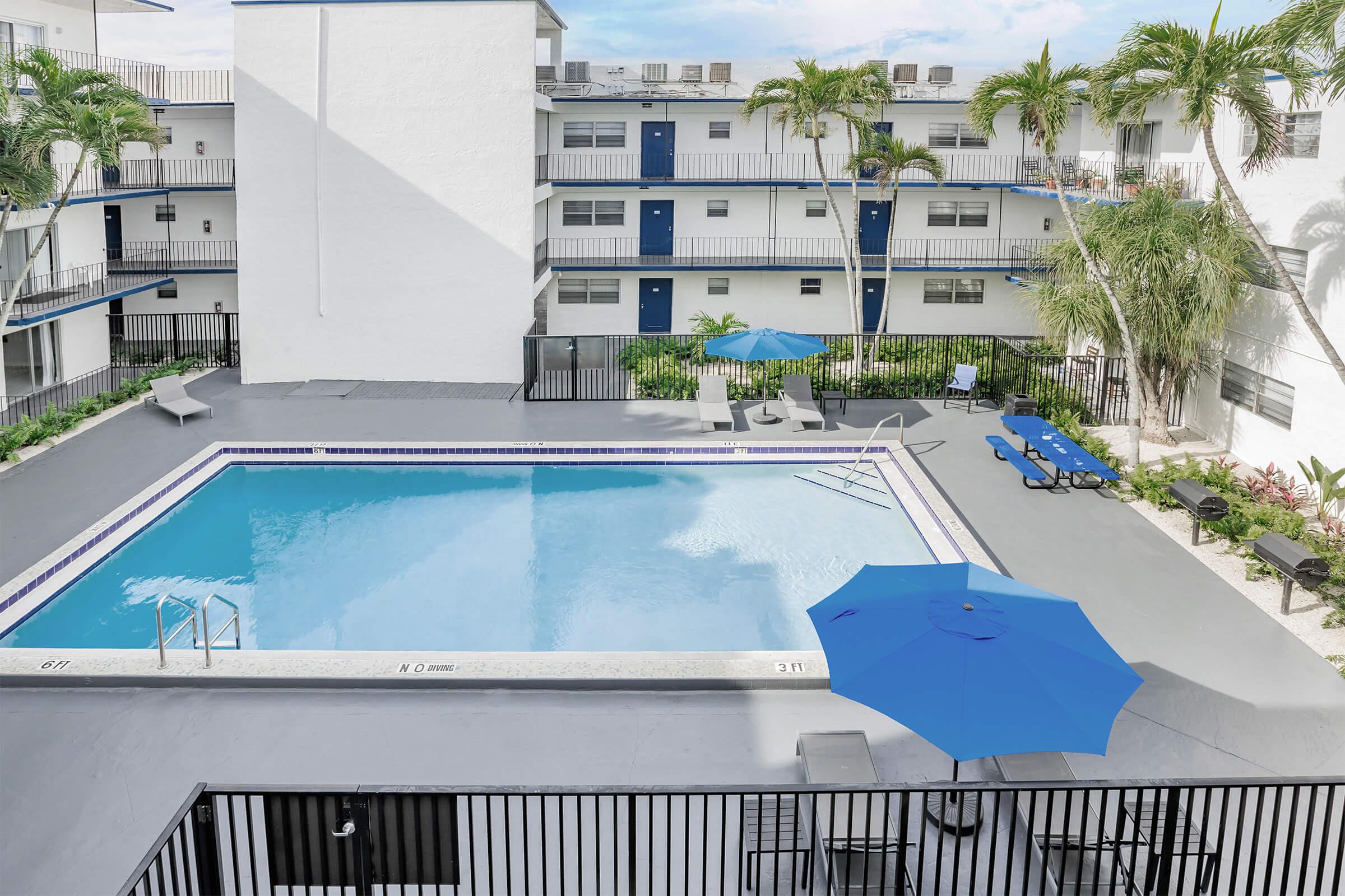 A clear swimming pool surrounded by a patio area with blue umbrellas and lounge chairs. The pool is in an apartment complex courtyard, featuring palm trees and well-maintained landscaping. The building has a modern design with multiple floors and balconies.