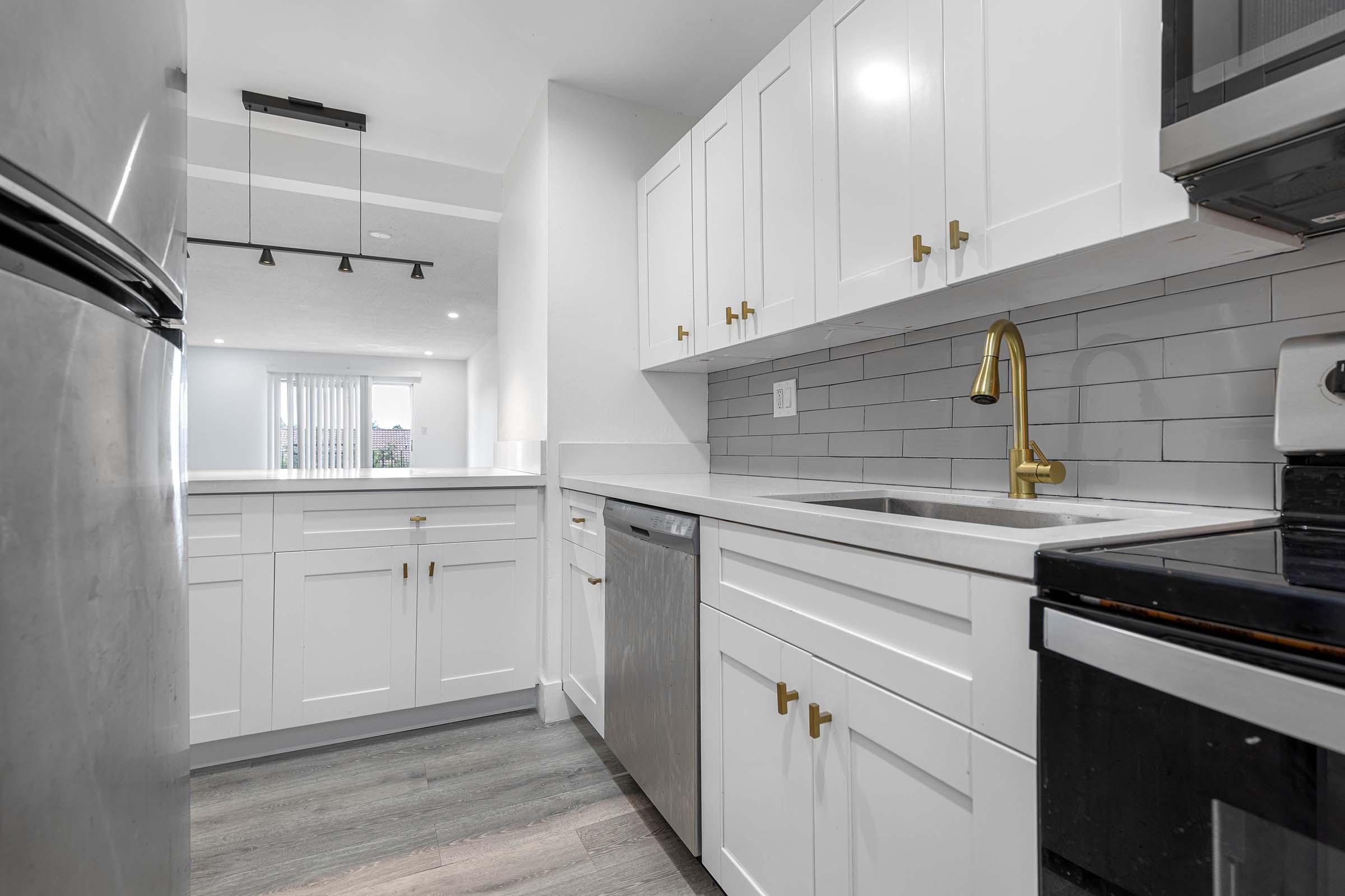 Modern kitchen featuring white cabinets with gold handles, a stainless steel refrigerator, and a sleek countertop. Includes a dishwasher and a black stove, with gray tiled backspash and wooden floors. A bright light fixture hangs from the ceiling, illuminating the space.