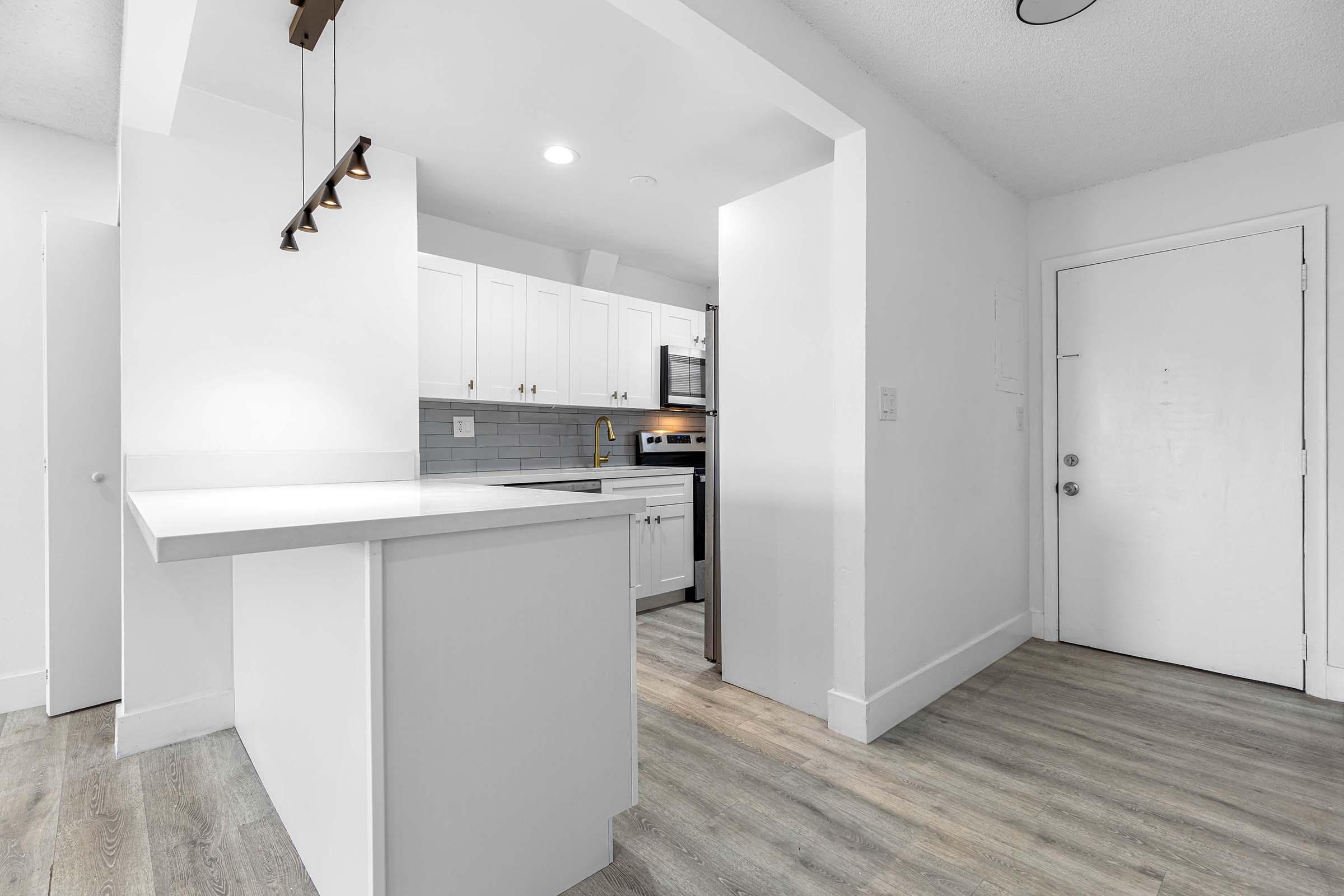 A bright kitchen area featuring white cabinetry, a modern stove, and a countertop with a small overhang. The adjacent space is open, leading to a white door, and the flooring is light-colored wood. Natural light flows in, creating an airy ambiance.