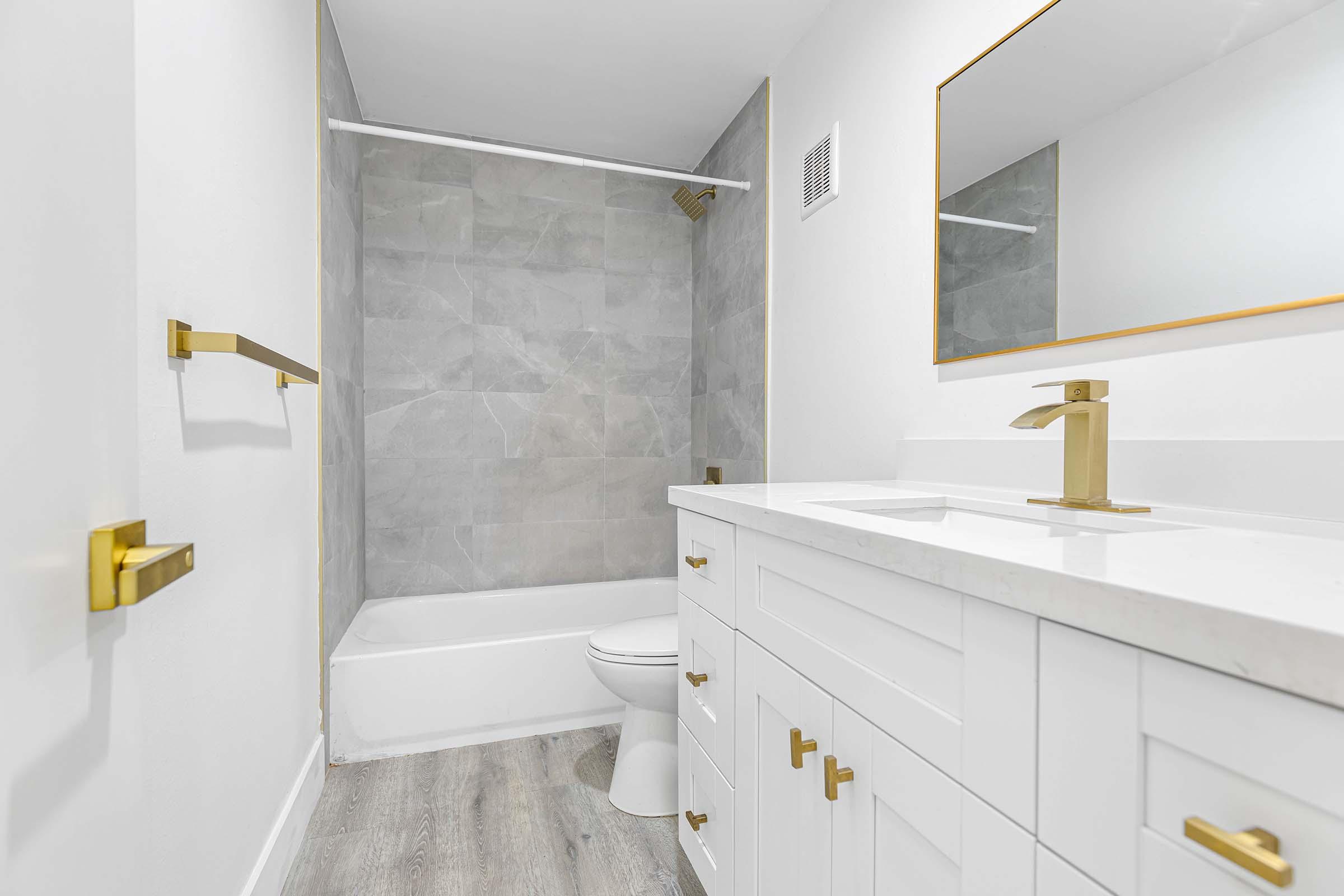 Modern bathroom featuring a white vanity with gold fixtures, a bathtub against a gray tiled wall, a large mirror, and light wood flooring. The walls are painted white, creating a bright and airy atmosphere.