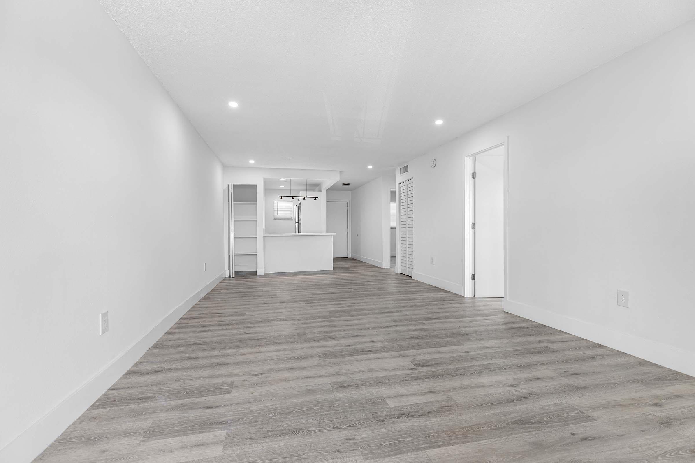 A spacious, empty room with light-colored walls and laminate flooring. On the left, there's a doorway leading to a kitchen area with a bar counter. The room features bright, recessed lighting and a second doorway on the right, suggesting a modern, minimalist design.