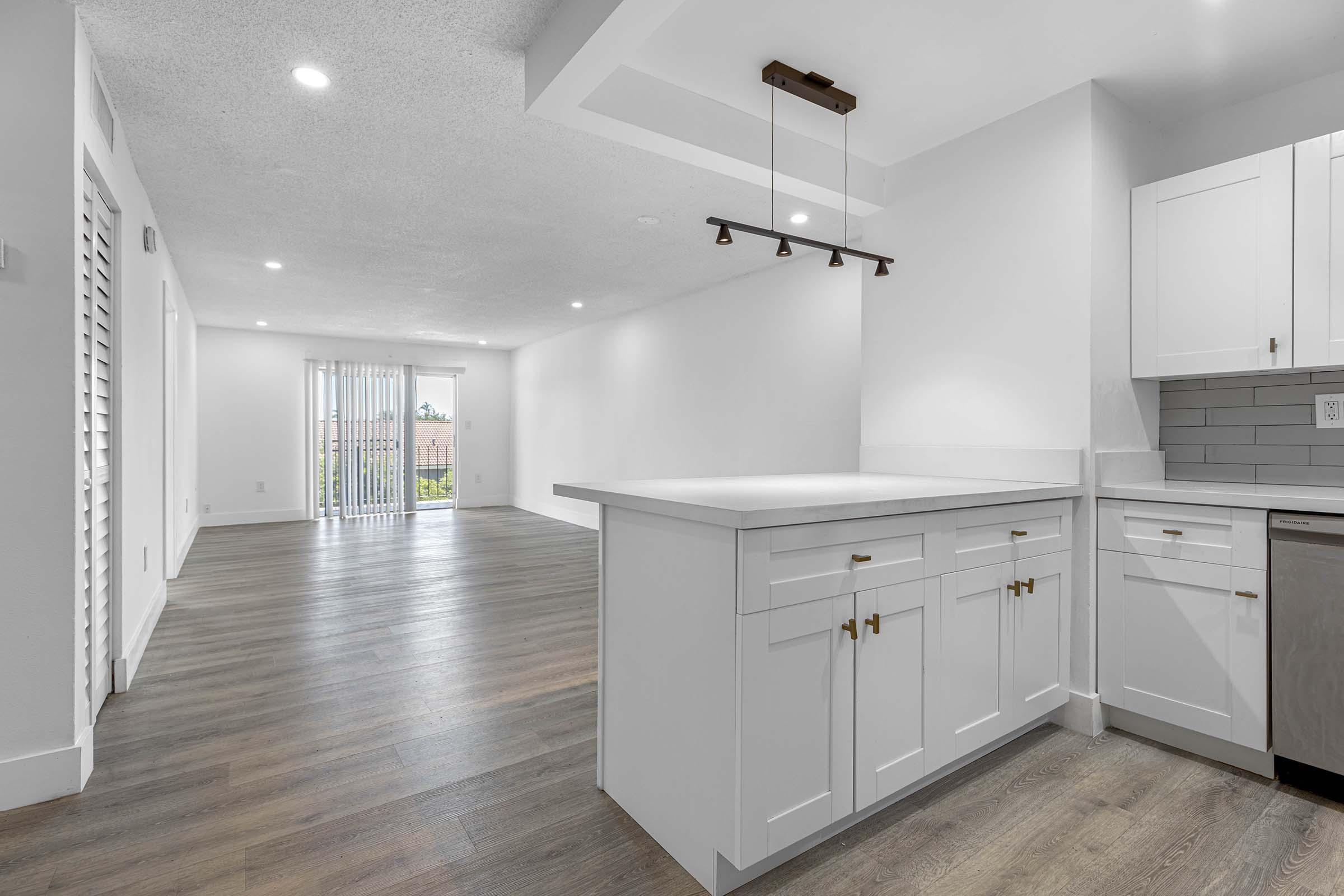 Modern kitchen and living area featuring wooden flooring, white cabinetry, and stainless steel appliances. The kitchen includes a countertop and a pendant light, while the living area is spacious with large sliding glass doors leading to a balcony. Natural light fills the room, enhancing the neutral color palette.