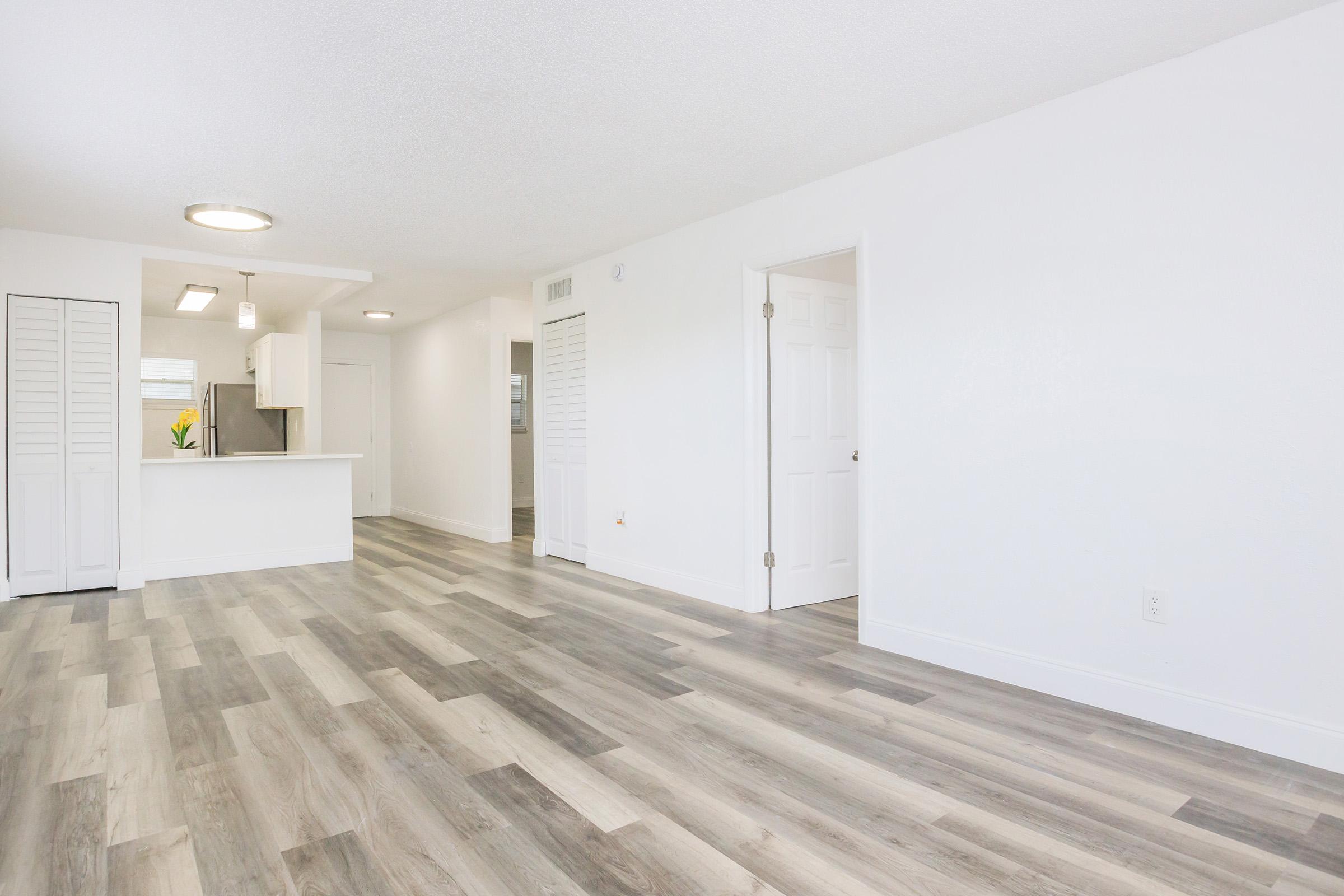 Spacious, modern interior of a bright, empty room featuring light wood-style flooring, white walls, and a small kitchen area visible in the background. The layout includes doors on the right and storage space on the left, creating an open and inviting atmosphere.