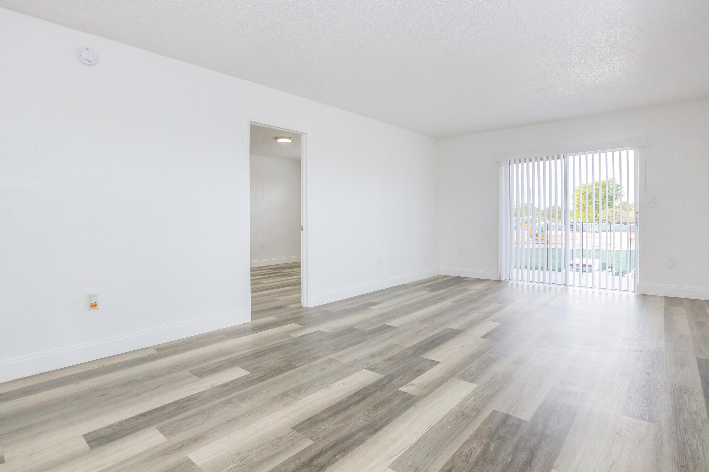 Spacious, empty room with light-colored walls and wooden flooring. A doorway leads to another room, and a sliding glass door with vertical blinds opens to a view outside. Natural light illuminates the space, creating a bright and airy atmosphere.