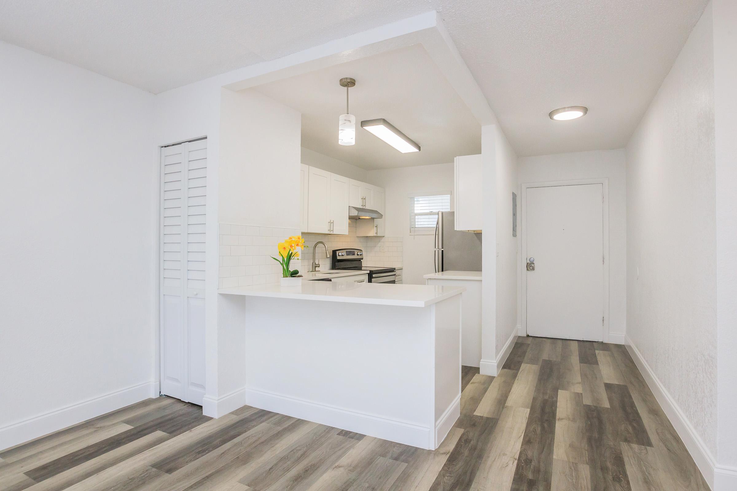 A modern kitchen featuring white cabinets, stainless steel appliances, and a small island. Bright lighting and a vase of yellow flowers add a cheerful touch. The floor has a stylish, wood-like finish, and there is an entryway with a white door.