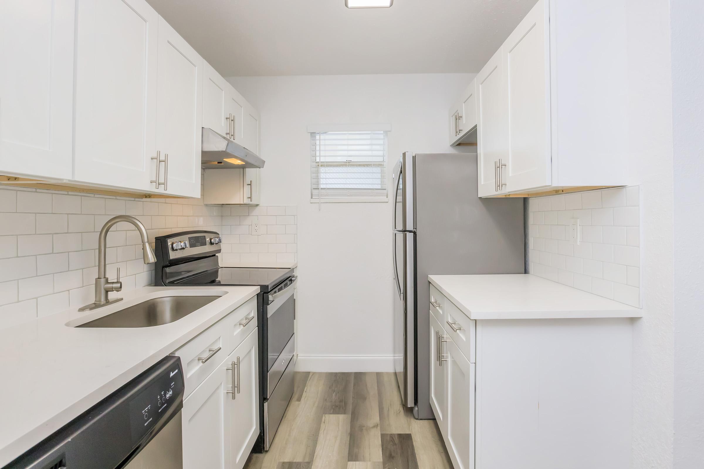 A modern kitchen featuring white cabinetry, stainless steel appliances, a double sink, and a window providing natural light. The design includes a combination of black and silver appliances and a light-colored countertop, creating a clean and contemporary look. The flooring is light wood-like laminate.