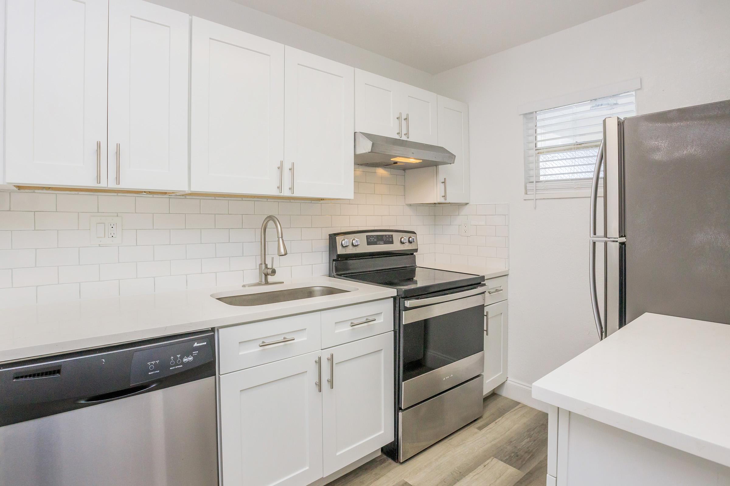 Modern kitchen featuring white cabinetry, a stainless steel stove and microwave, a stainless steel dishwasher, and a sleek refrigerator. The countertop is light-colored, and the backsplash consists of white subway tiles, complemented by natural light from a window.