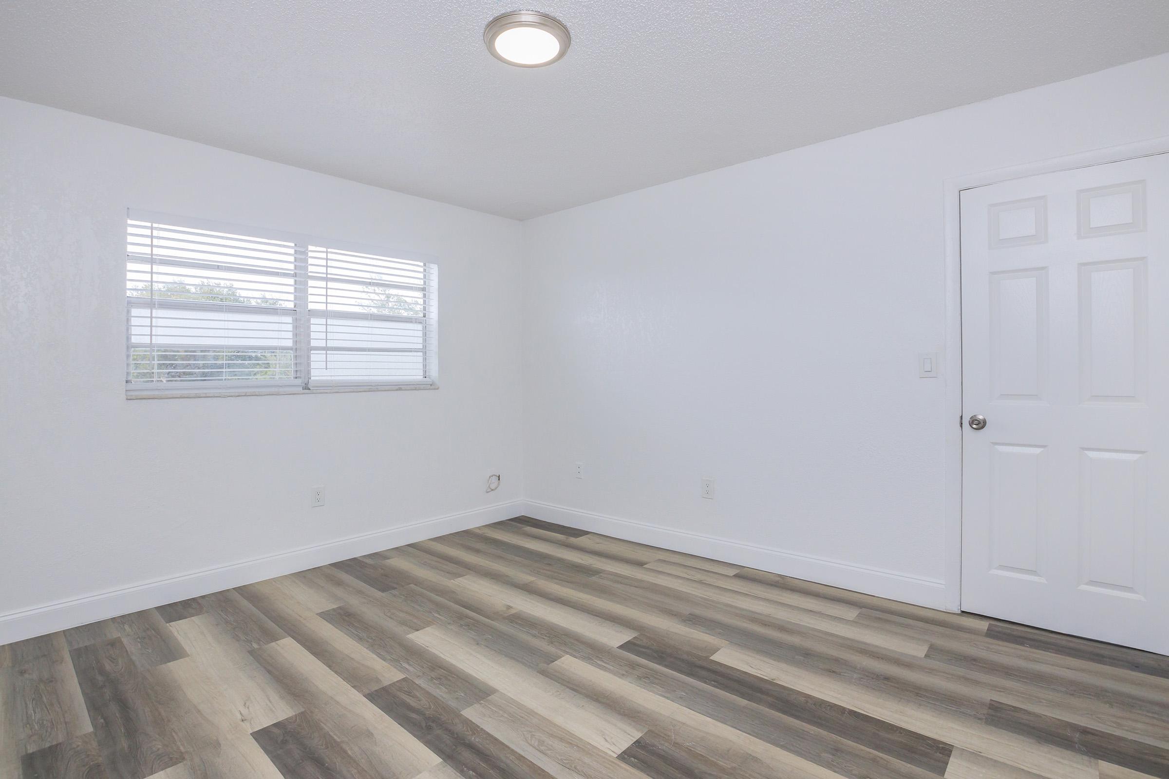 Empty room with light-colored walls and a window featuring blinds. The floor is covered in light wood-like laminate, and there is a door on the right side of the image. The space is well-lit and appears clean and newly updated.