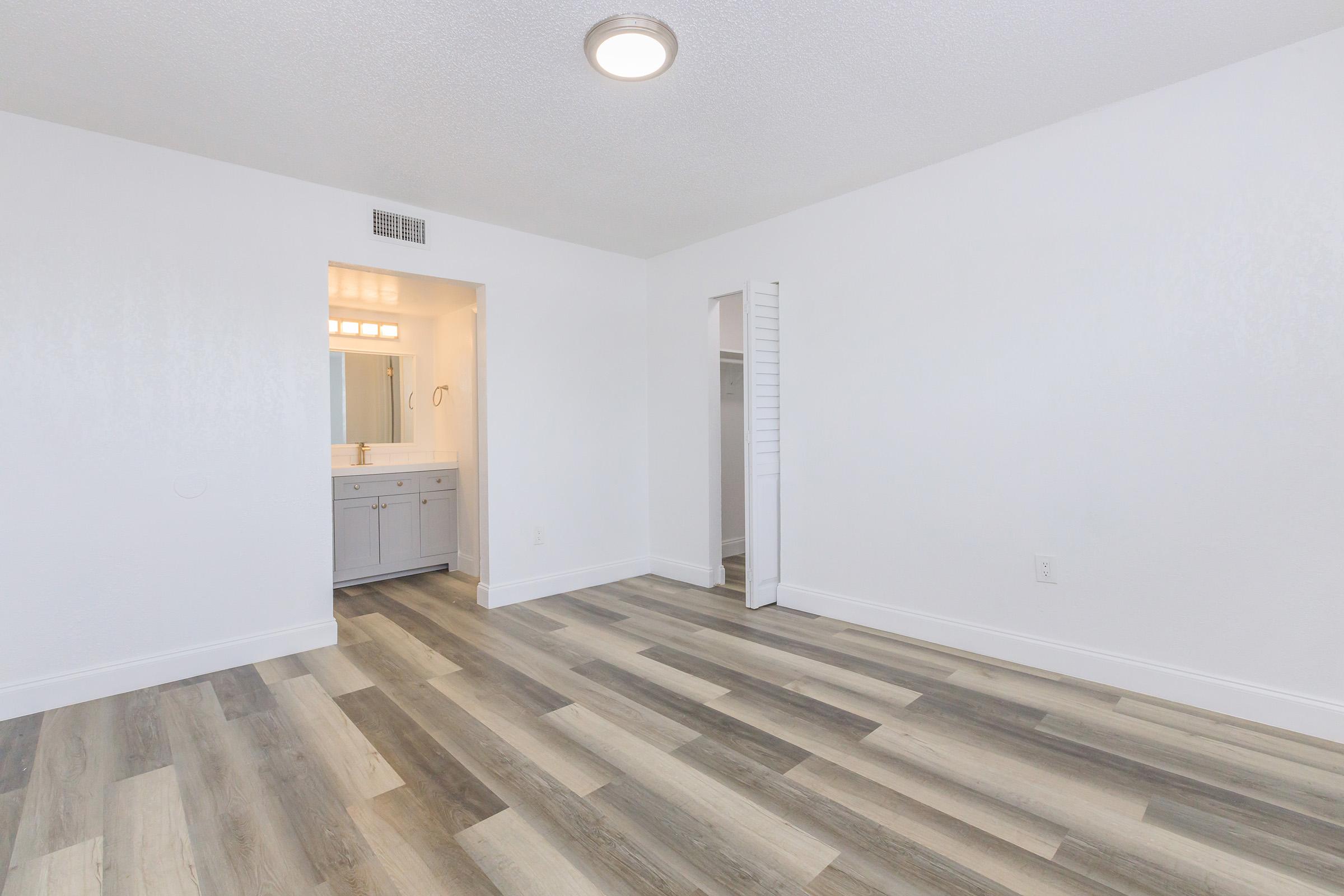 A spacious and well-lit room featuring light-colored walls and a wooden floor. There is a doorway leading to a bathroom with a vanity, and another closet space visible on the side. The overall ambiance is fresh and modern, perfect for various furniture arrangements.