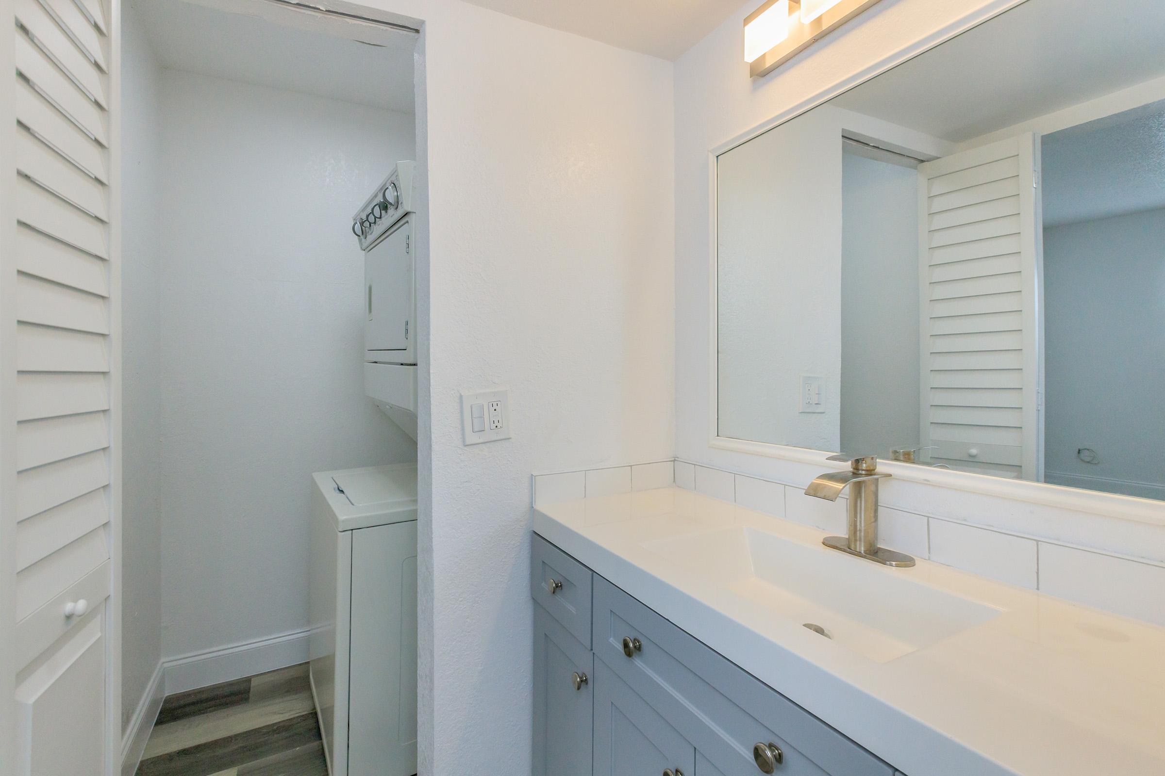 A modern bathroom featuring a white sink with a sleek faucet, gray cabinetry, and a large mirror. To the side, there is a small laundry area with a stacked washer and dryer, complemented by white shutter doors. The walls are painted white, and the floor has a stylish tile design.