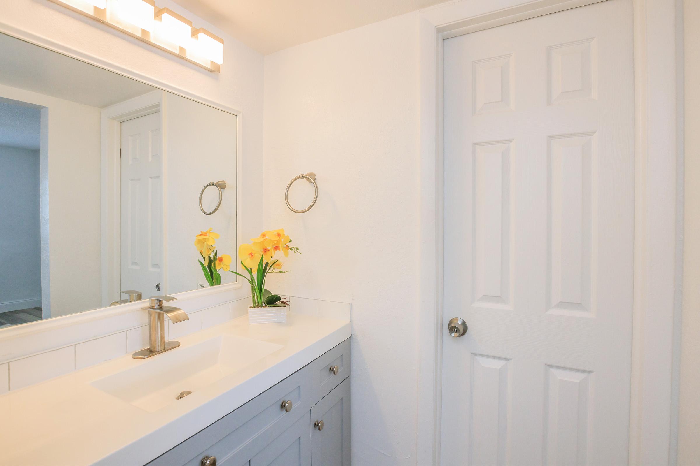 A bright, modern bathroom featuring a rectangular sink with a sleek faucet, a large mirror above, and a white cabinet below. Yellow flowers in a small vase add a pop of color. The walls are painted white, and a closed door is visible in the background. Soft lighting enhances the clean, fresh atmosphere.